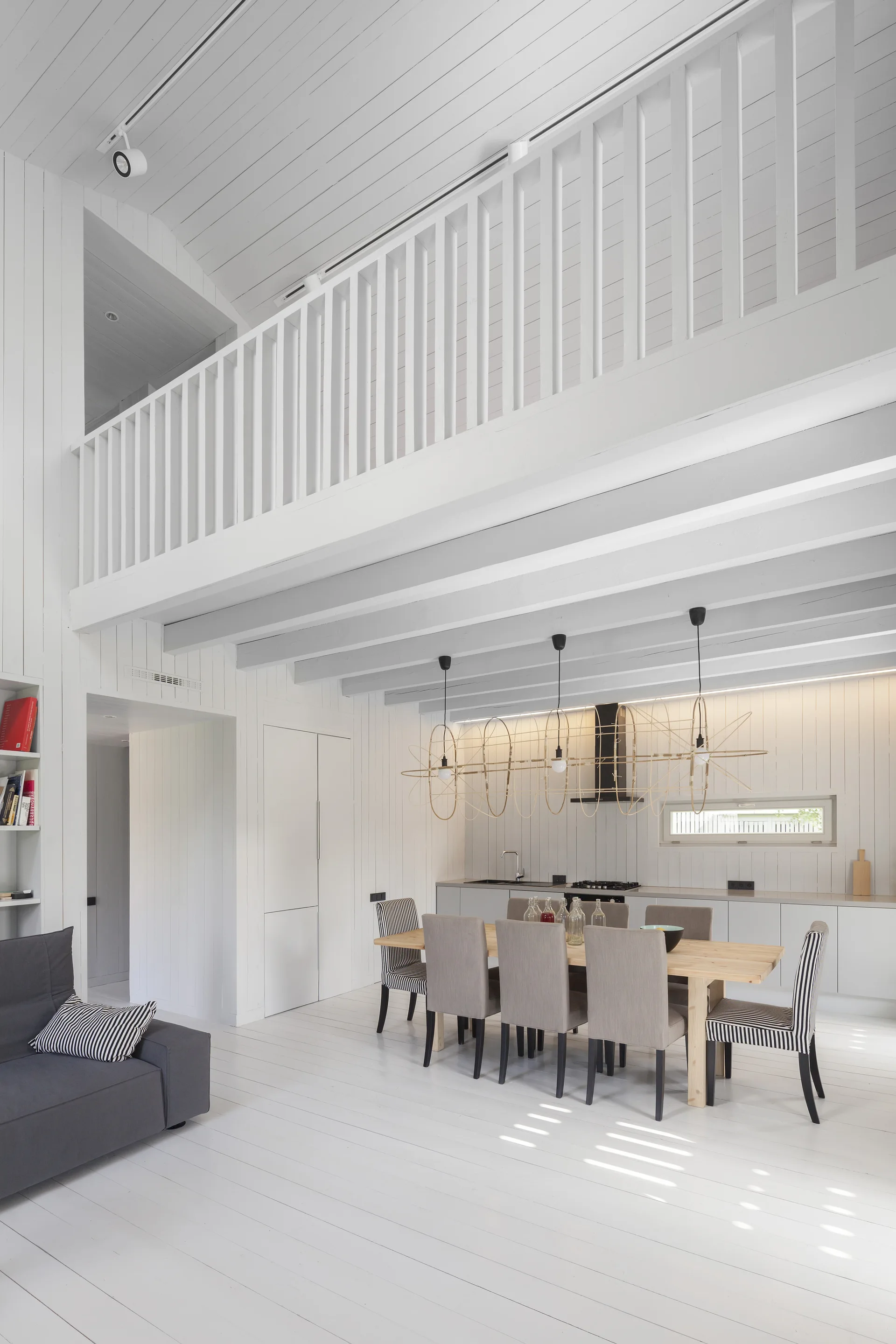 A bright white dining area with a wooden table, gray and striped chairs, a wireframe pendant light, and a loft mezzanine above.