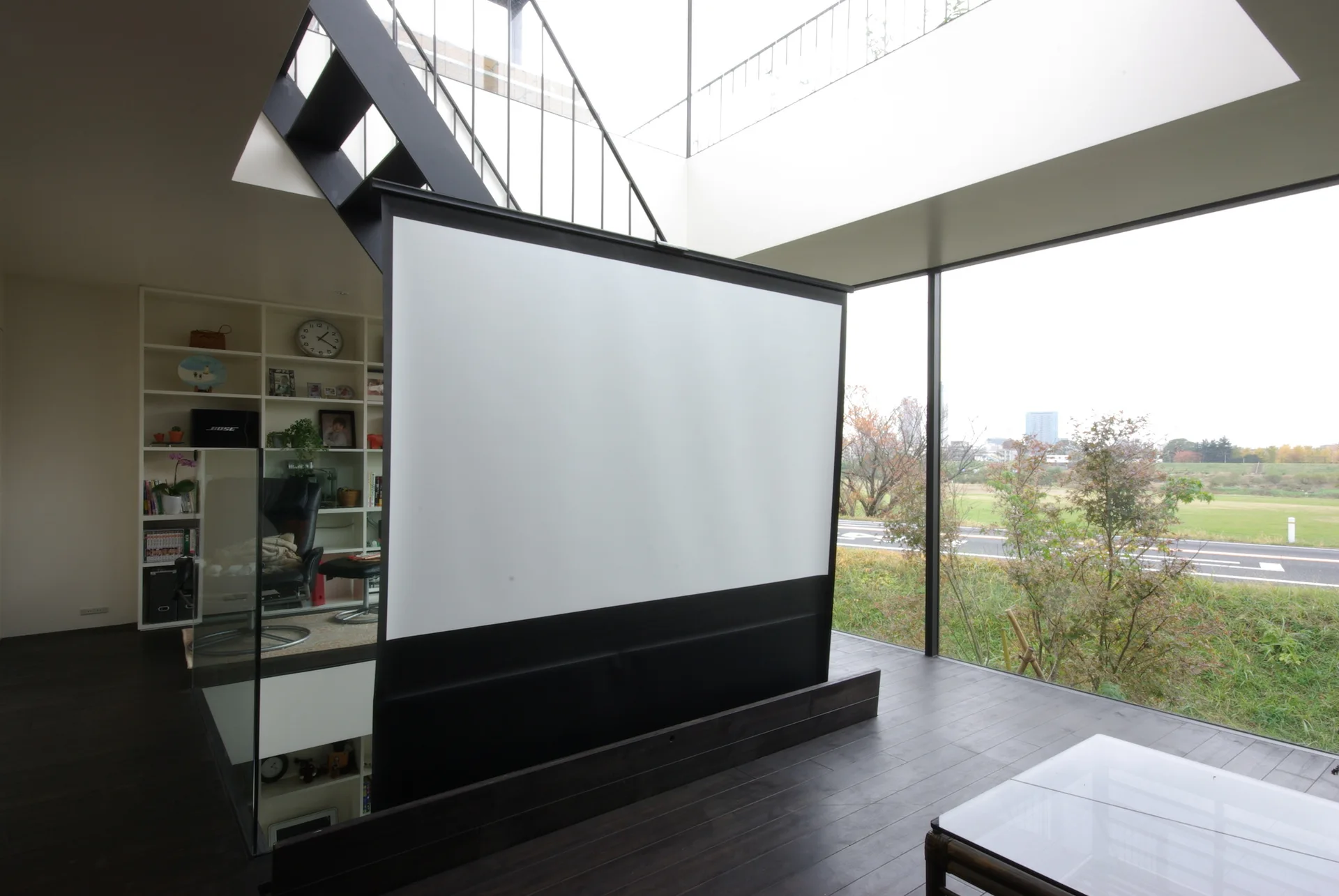 A modern living space features a large white projection screen, dark wood floors, a black metal staircase, and a large glass window wall.