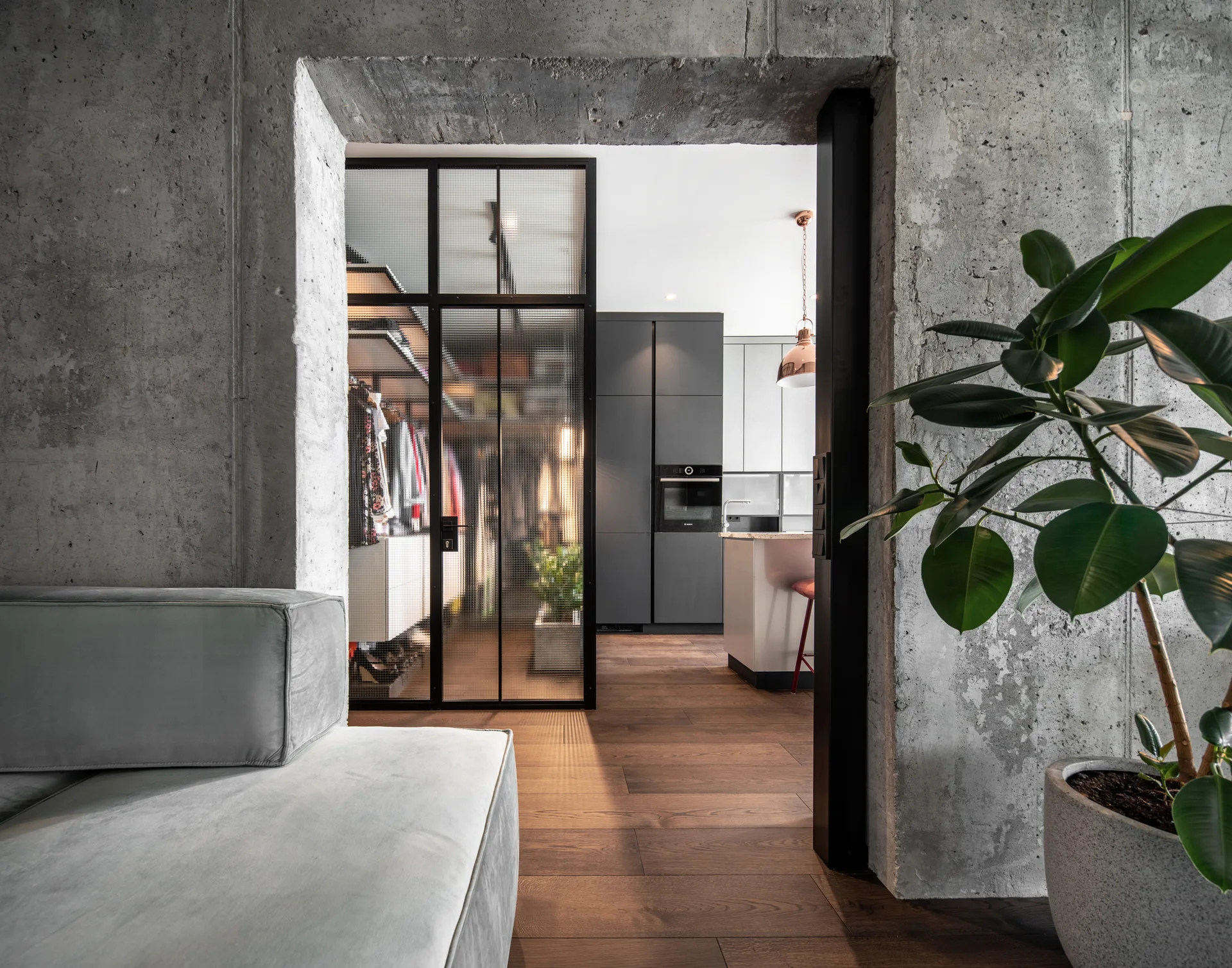 View from a living room with raw concrete walls and a grey sofa, looking toward a black-framed glass closet partition and a modern kitchen.
