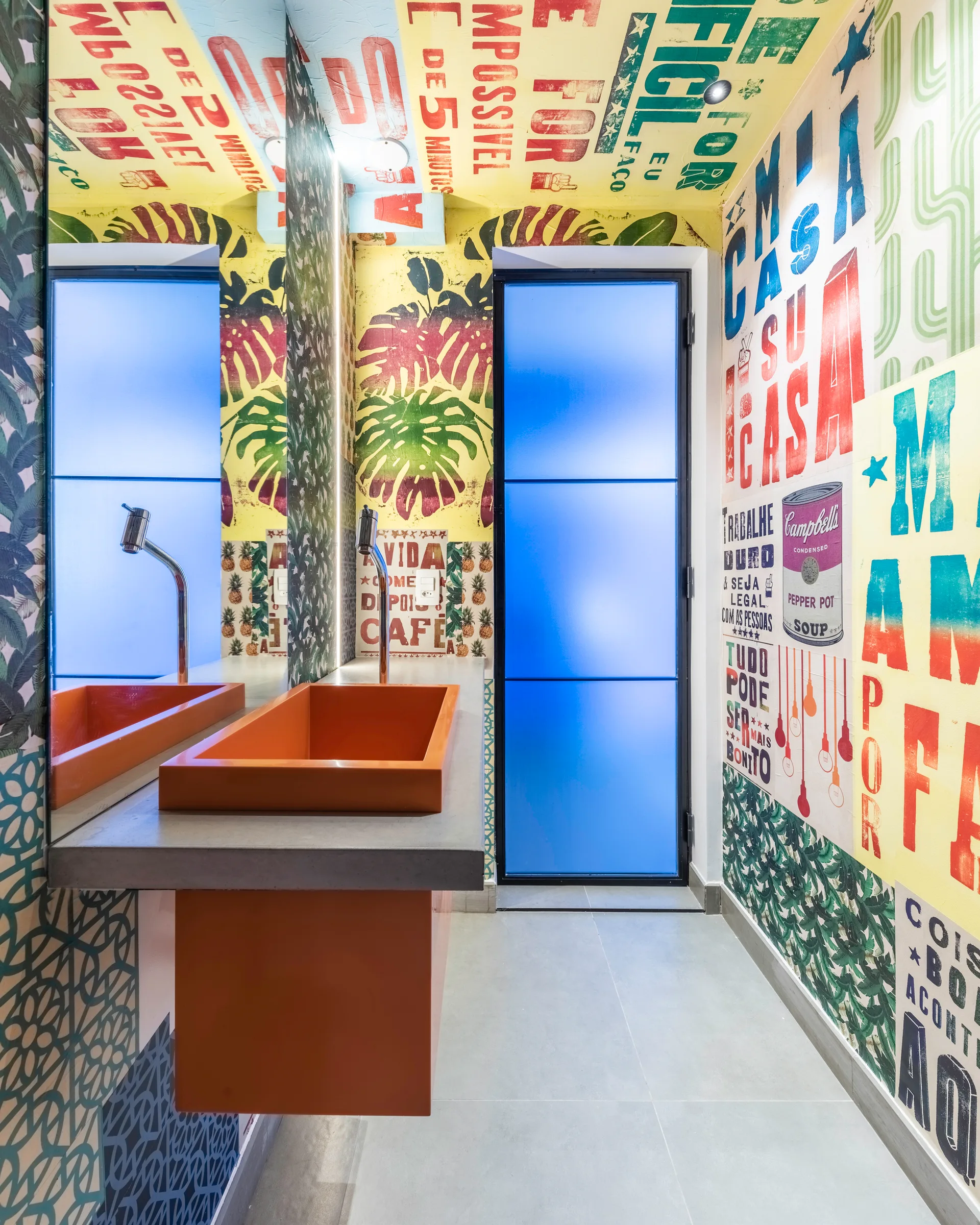 A powder room features colorful typography wallpaper, a wall-mounted concrete vanity with an orange rectangular basin, and a frosted blue glass door.
