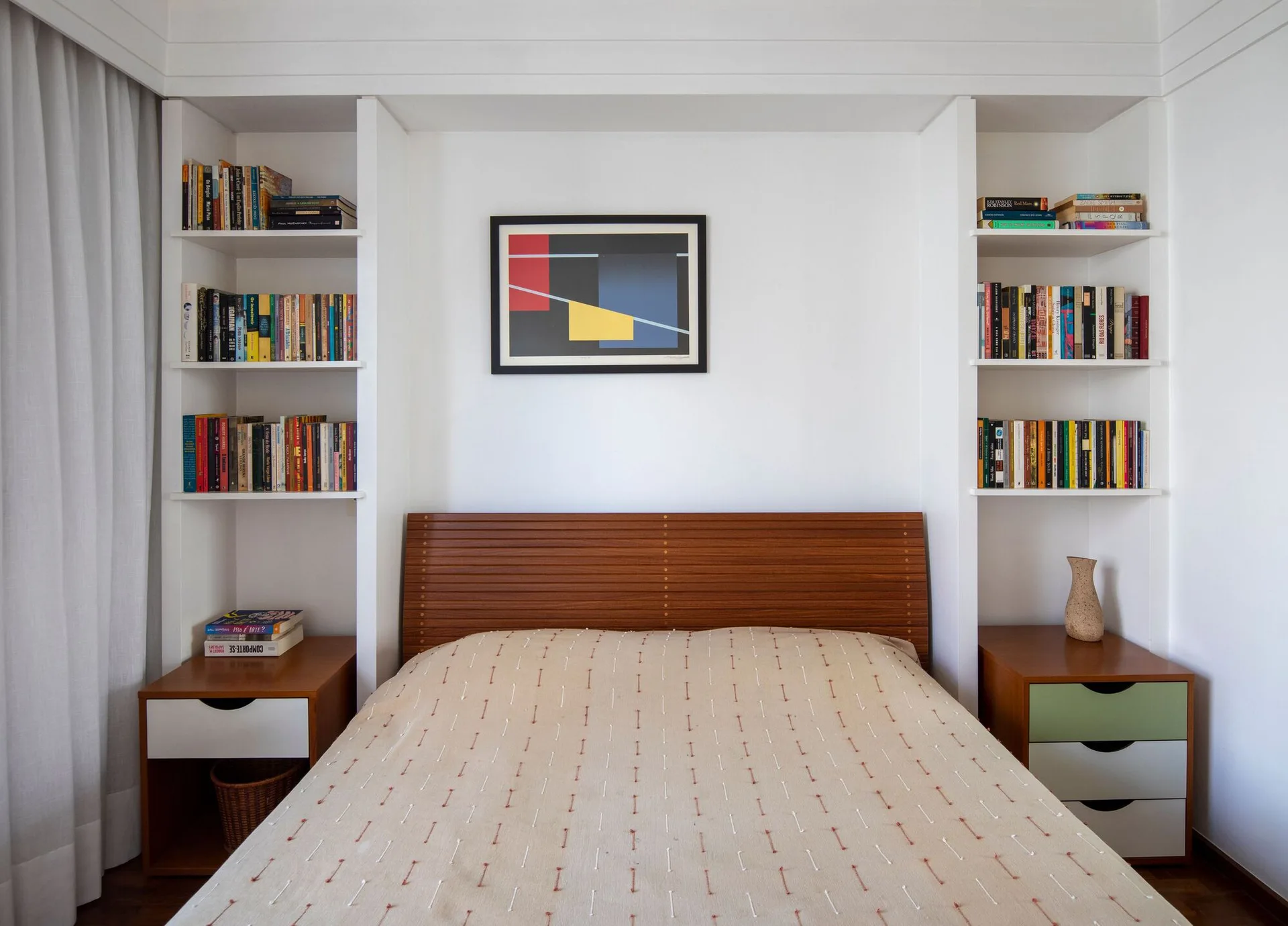 A bed with a wooden slatted headboard sits inside a white built-in alcove flanked by bookshelves and matching wood nightstands.