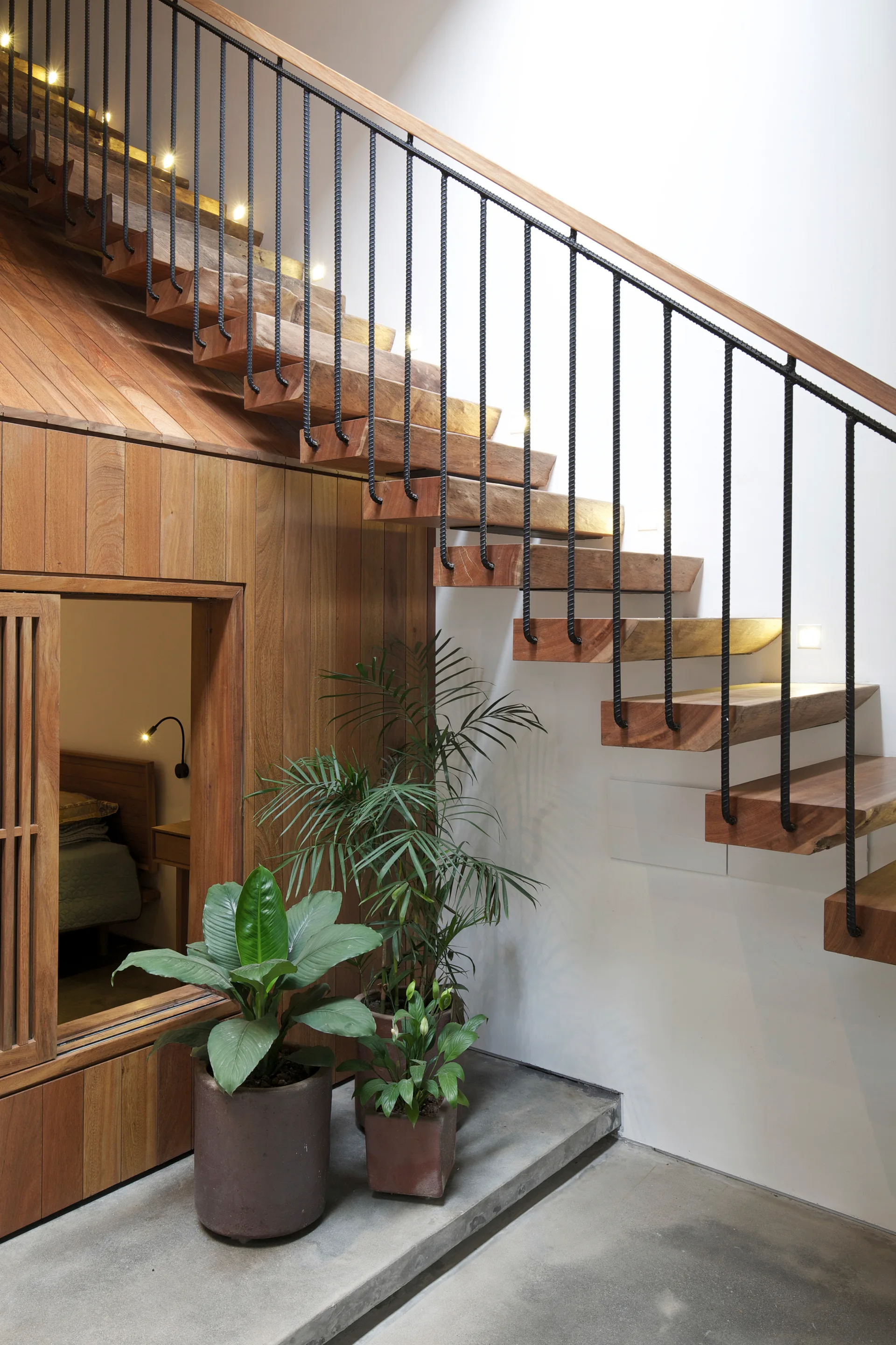 A floating wooden staircase with black iron spindles and integrated wall lighting stands above a polished concrete floor with potted plants.