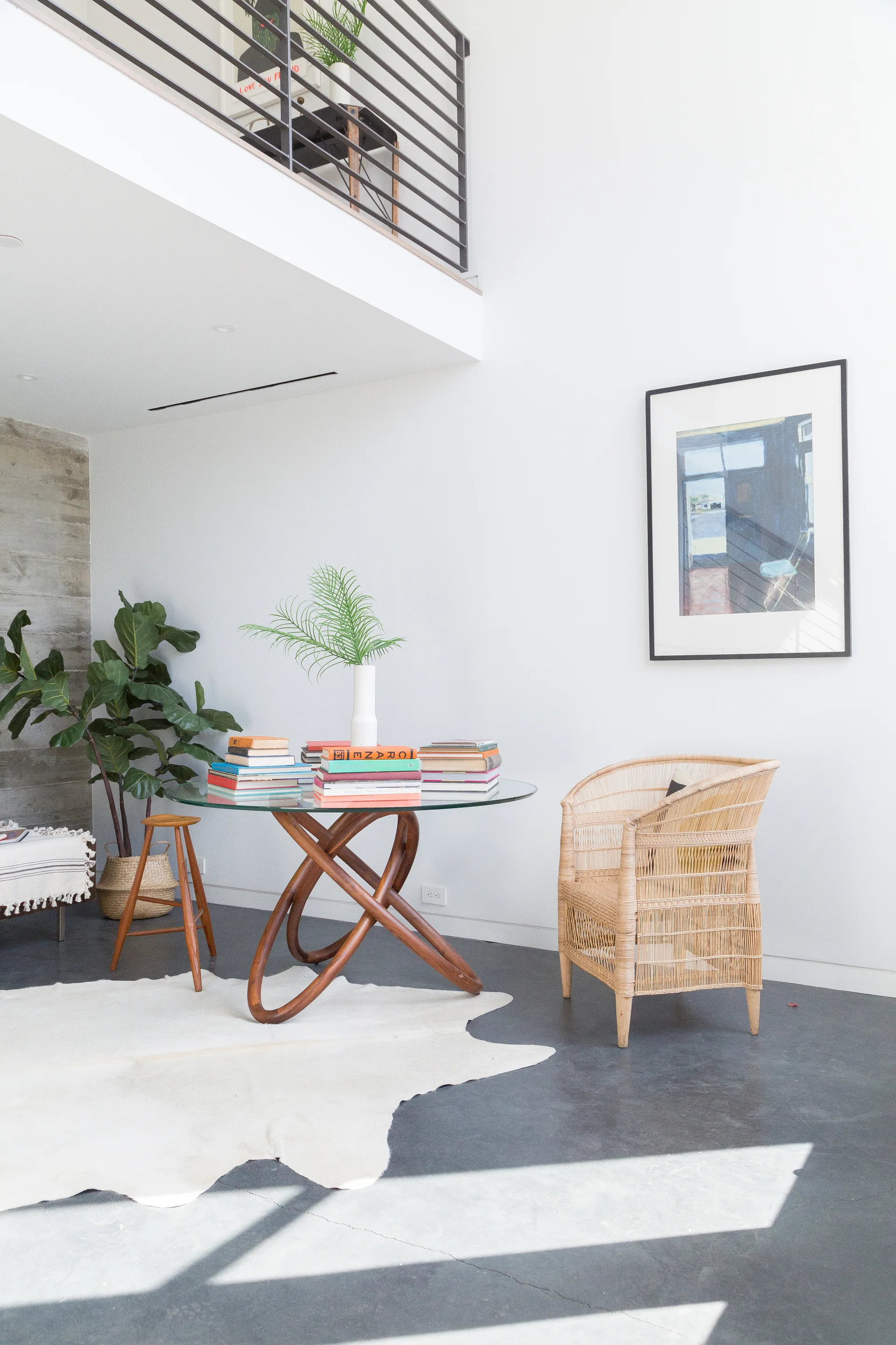 A light-filled dining space features a glass-top table with a looped wood base, a rattan armchair, and a white hide rug on concrete floors.