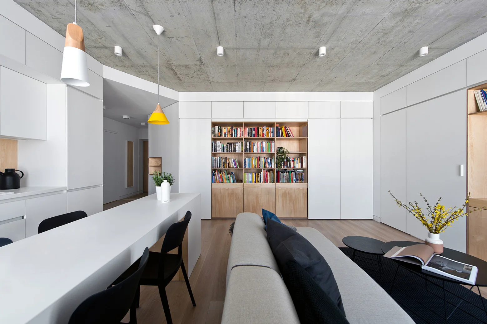 A modern open-plan living room features an exposed concrete ceiling, seamless white built-in storage, a recessed wooden bookcase, and a gray sofa.