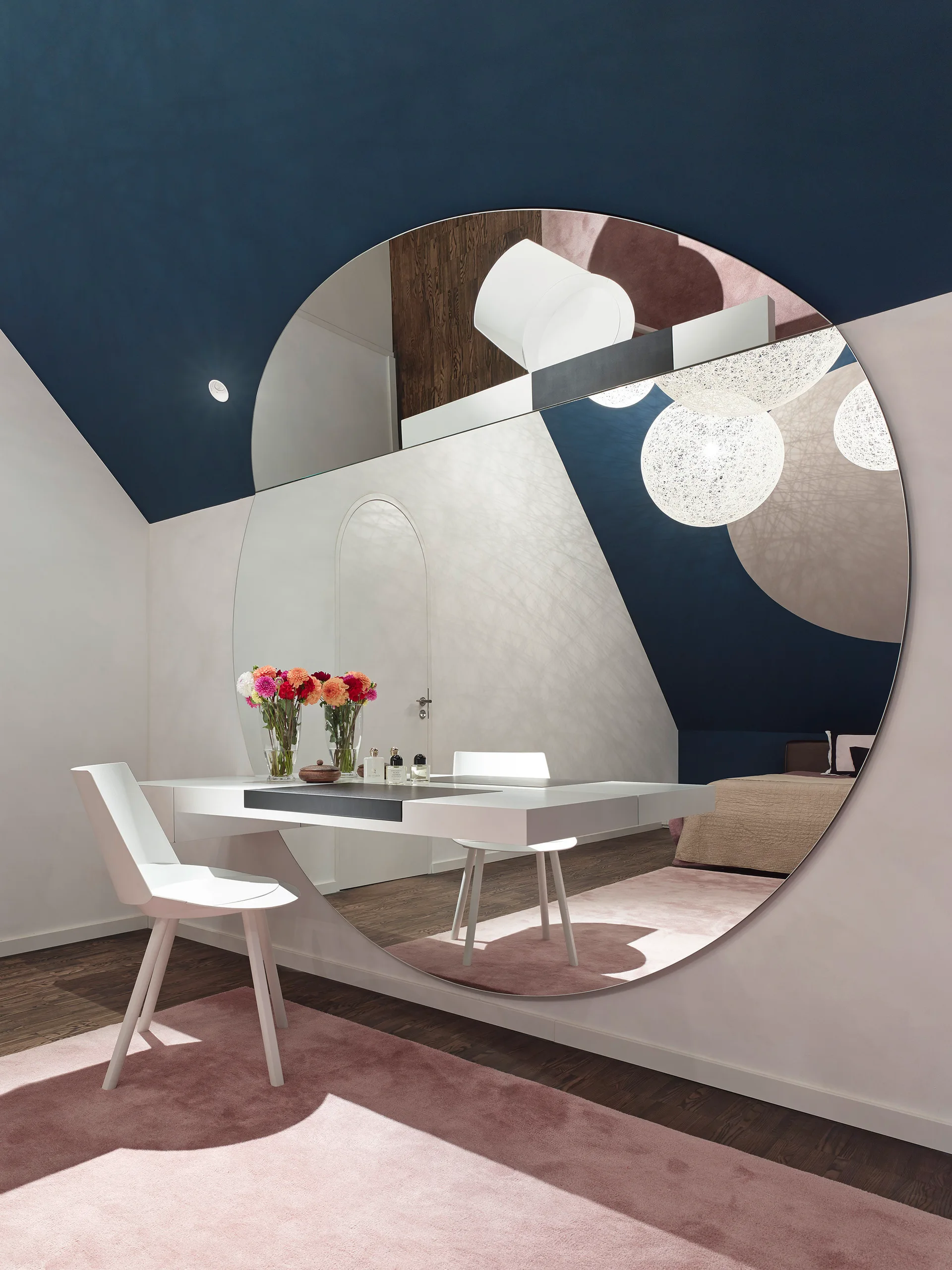 A white vanity desk and chair are set against a large round mirror reflecting spherical pendant lights and a deep blue ceiling.