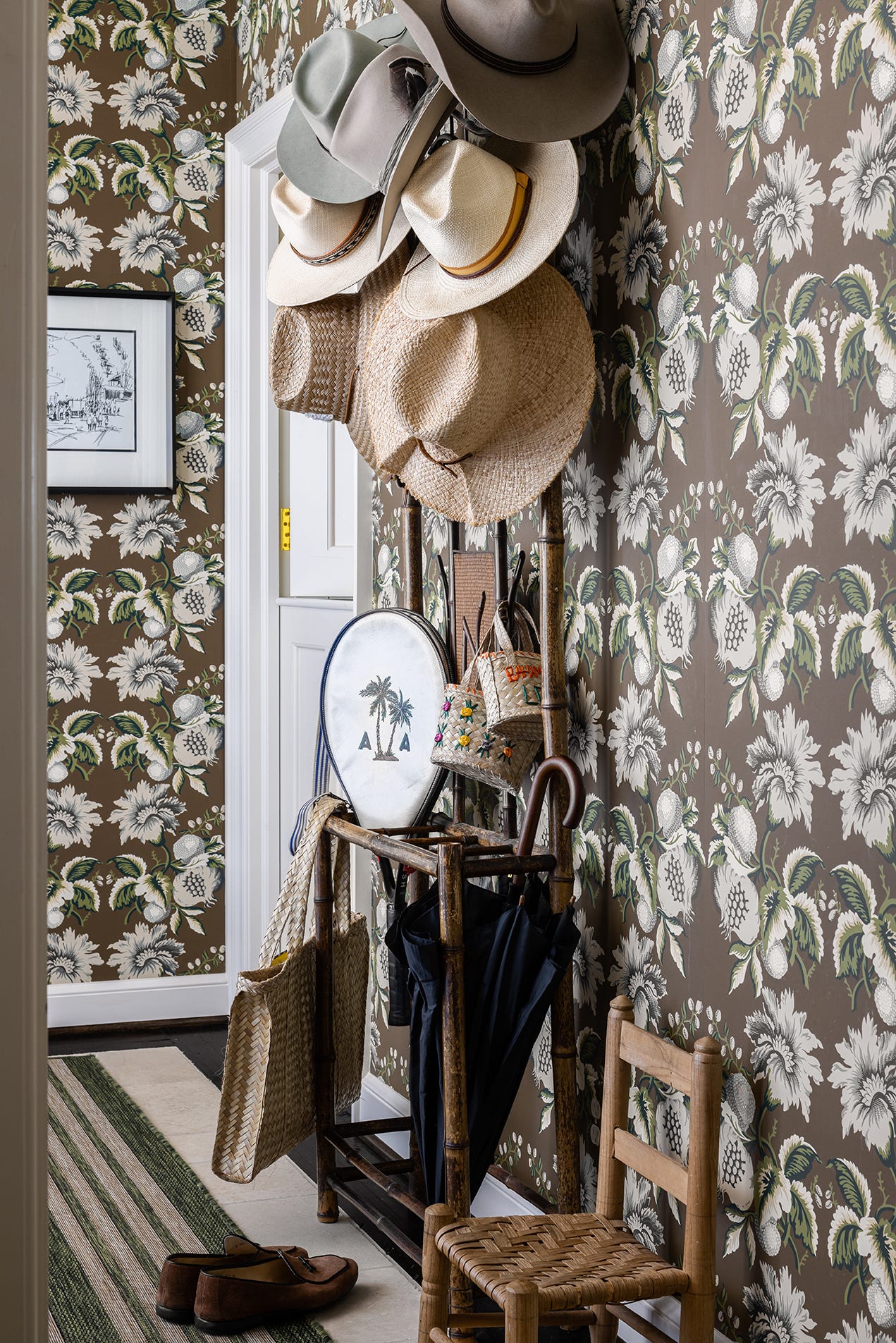 An entryway features brown botanical wallpaper, a bamboo hat rack filled with straw hats, a striped rug, and a small woven chair.