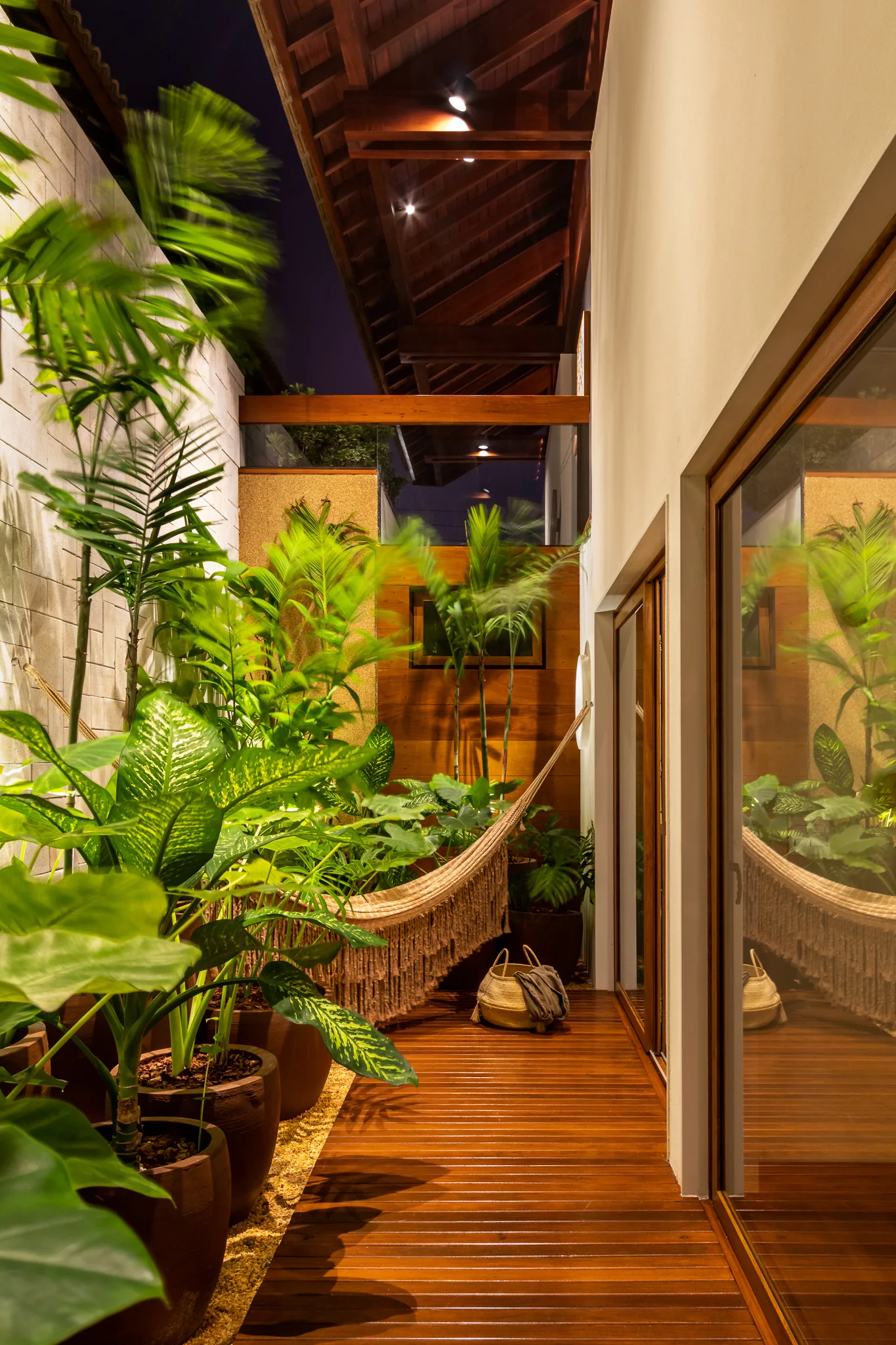 A narrow wooden deck is lined with large potted tropical plants and features a fringed woven hammock suspended beneath exposed wood ceiling beams at night.