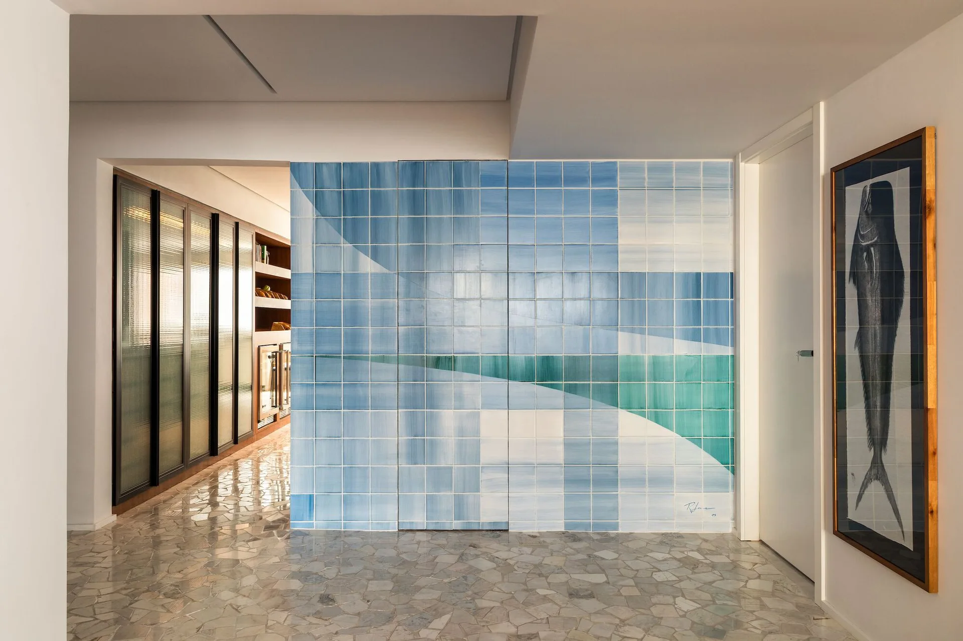A contemporary hallway features a prominent wall of blue and turquoise geometric tiles, paired with polished grey terrazzo flooring and glass cabinets.