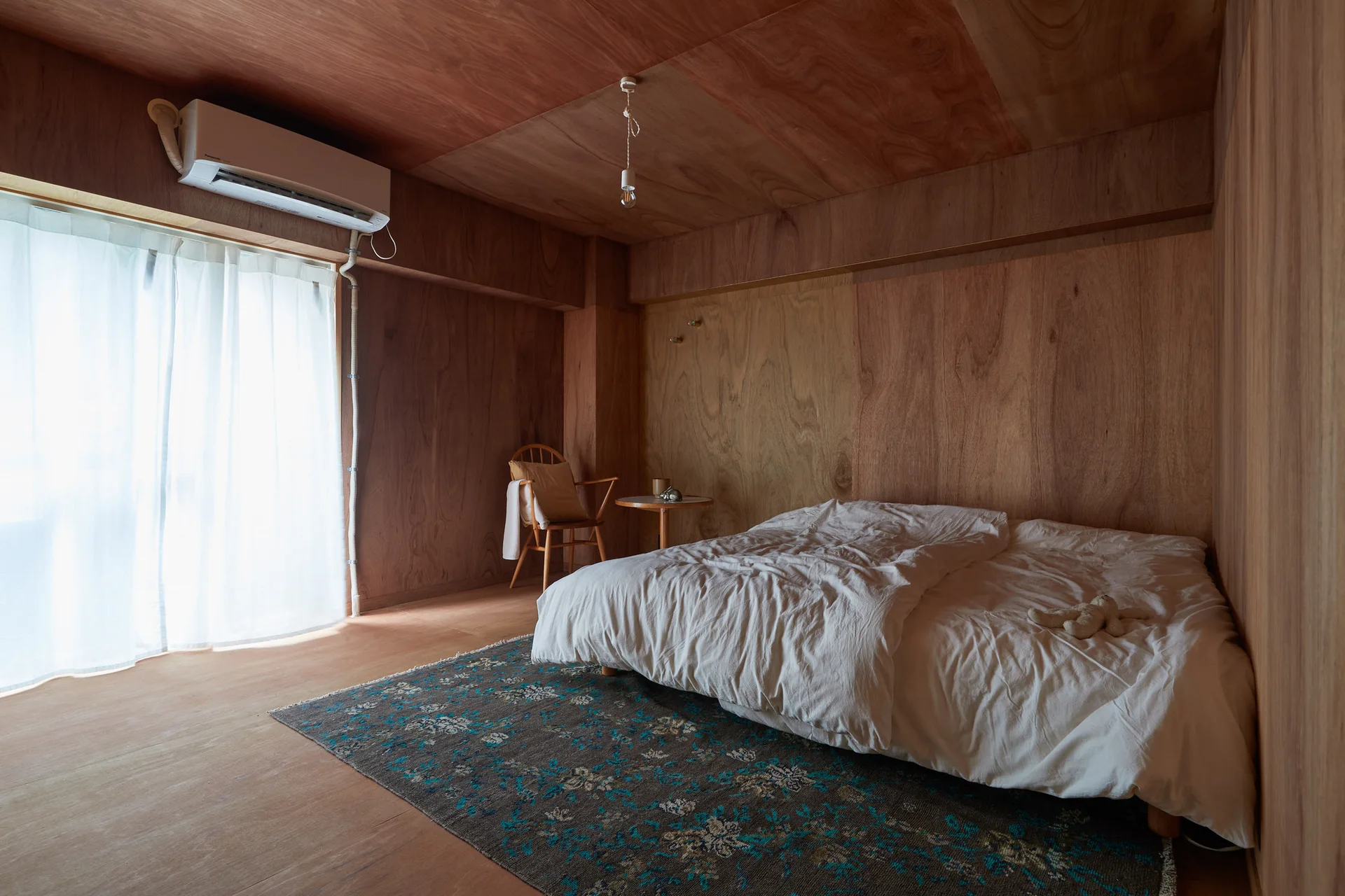 A minimalist bedroom featuring warm plywood paneled walls and ceiling, a low bed with white bedding, and a dark patterned rug.