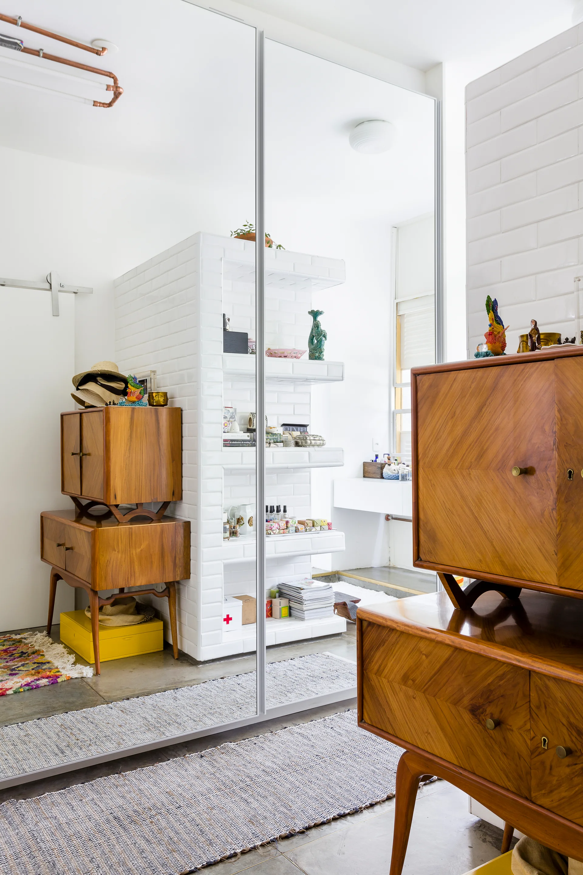 Warm wood mid-century cabinets stand against a large mirrored wall next to white tiled built-in shelving on a concrete floor.