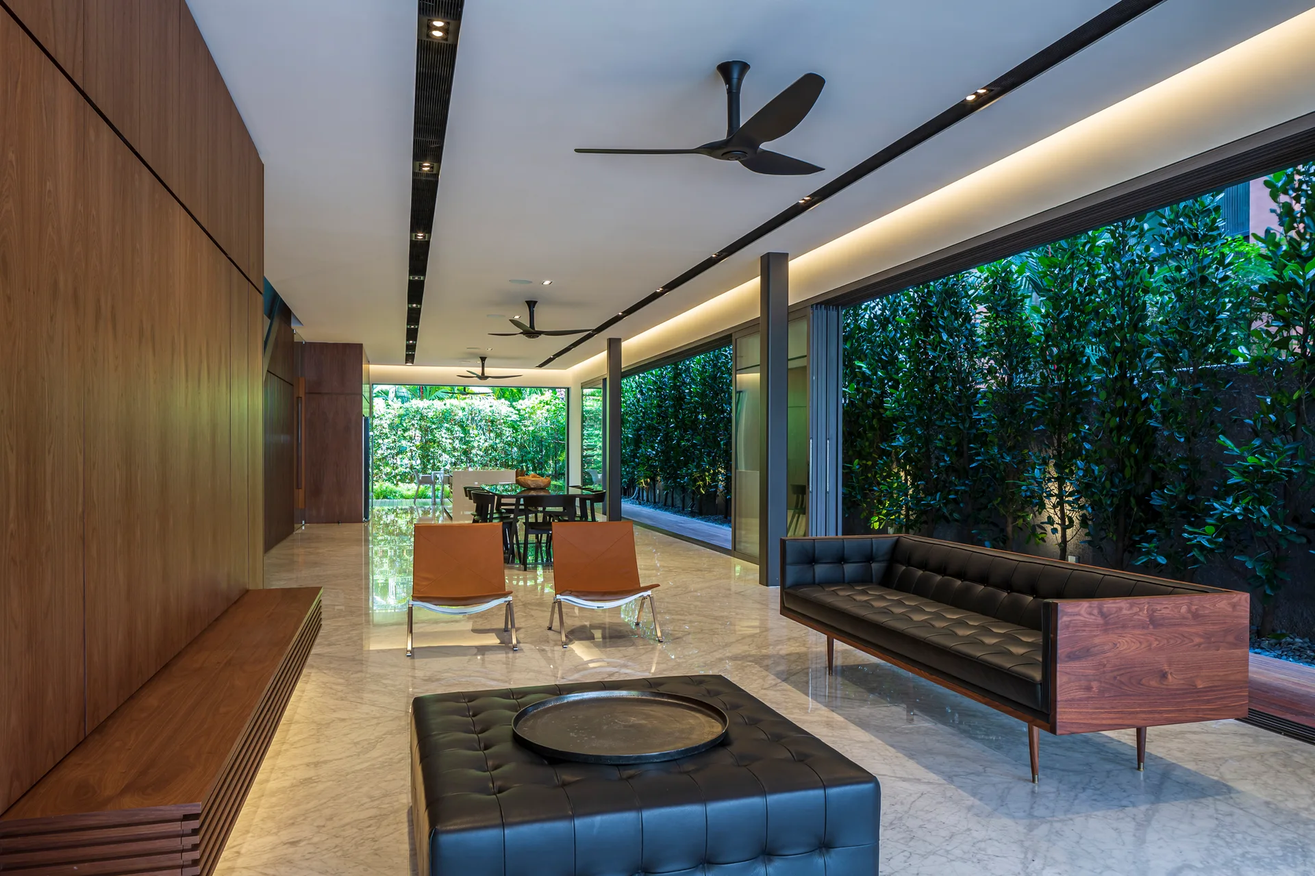 A modern open-concept living room with polished marble floors, black leather seating, warm wood wall paneling, and glass doors opening to a lush garden.