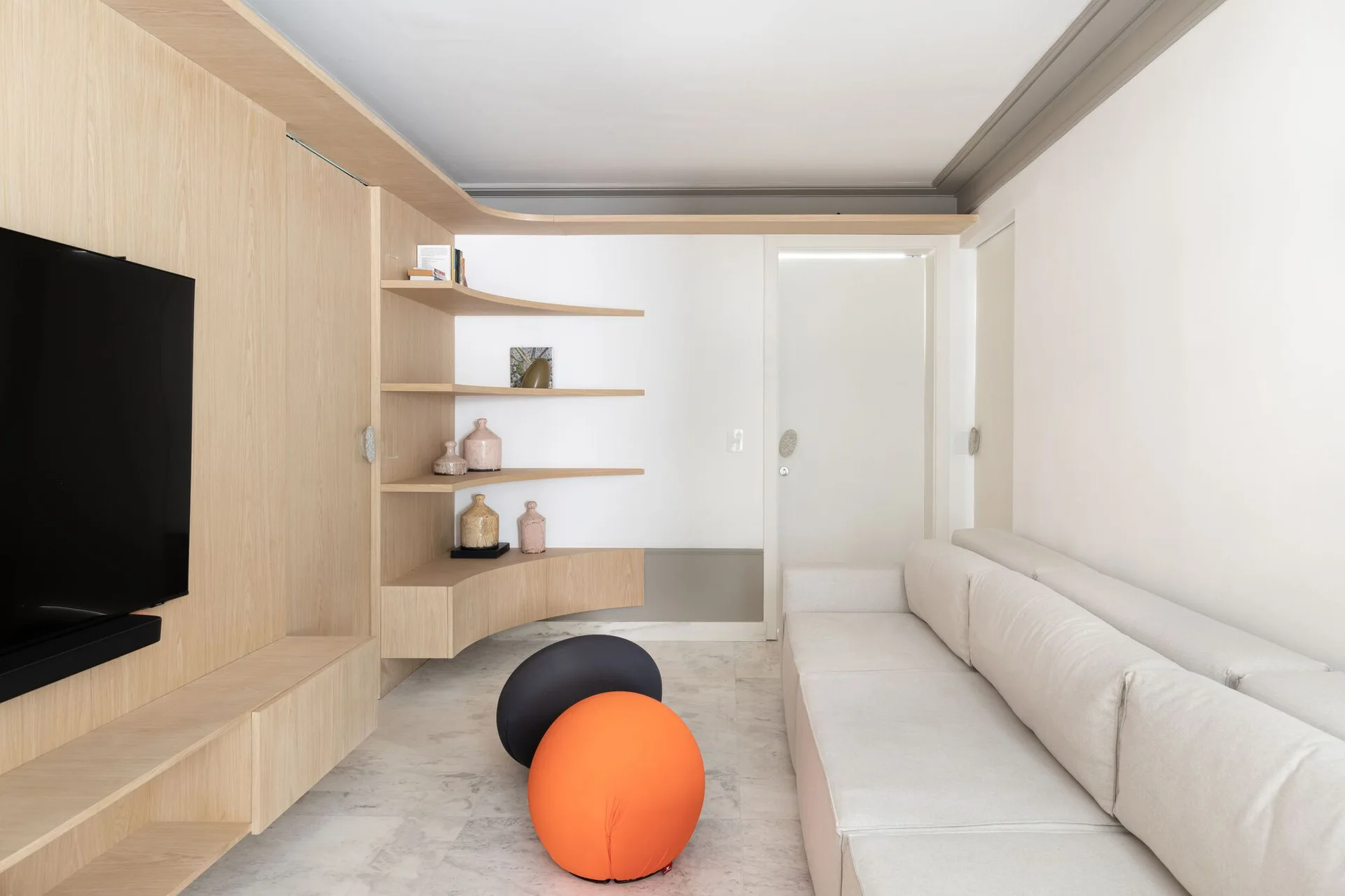 A minimalist living room features a custom light wood TV unit with curved corner shelving, a white sofa, and two spherical floor cushions.