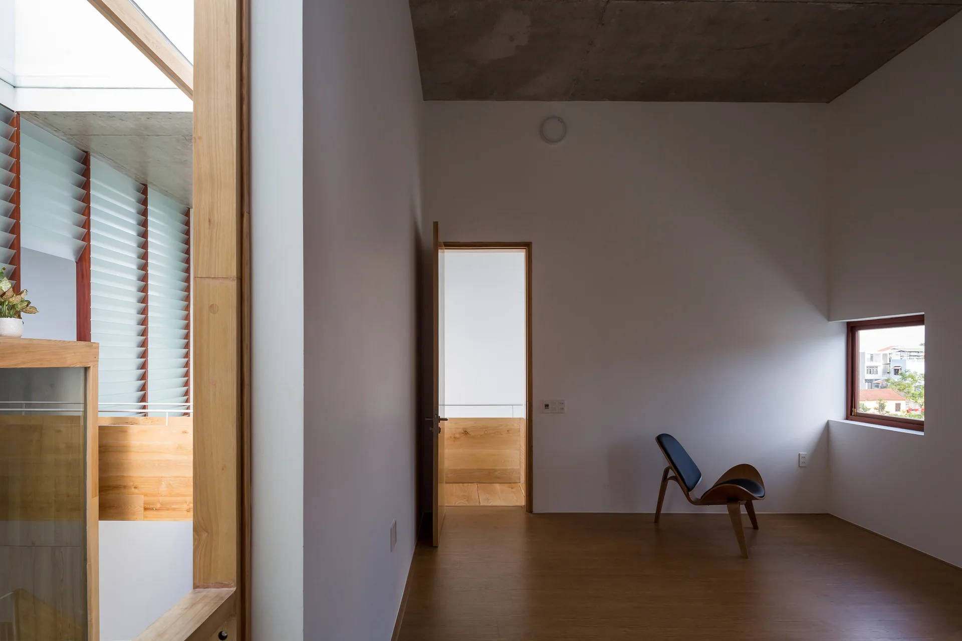 A minimalist room features white walls, warm wood flooring, an exposed concrete ceiling, and a single modern wooden lounge chair.