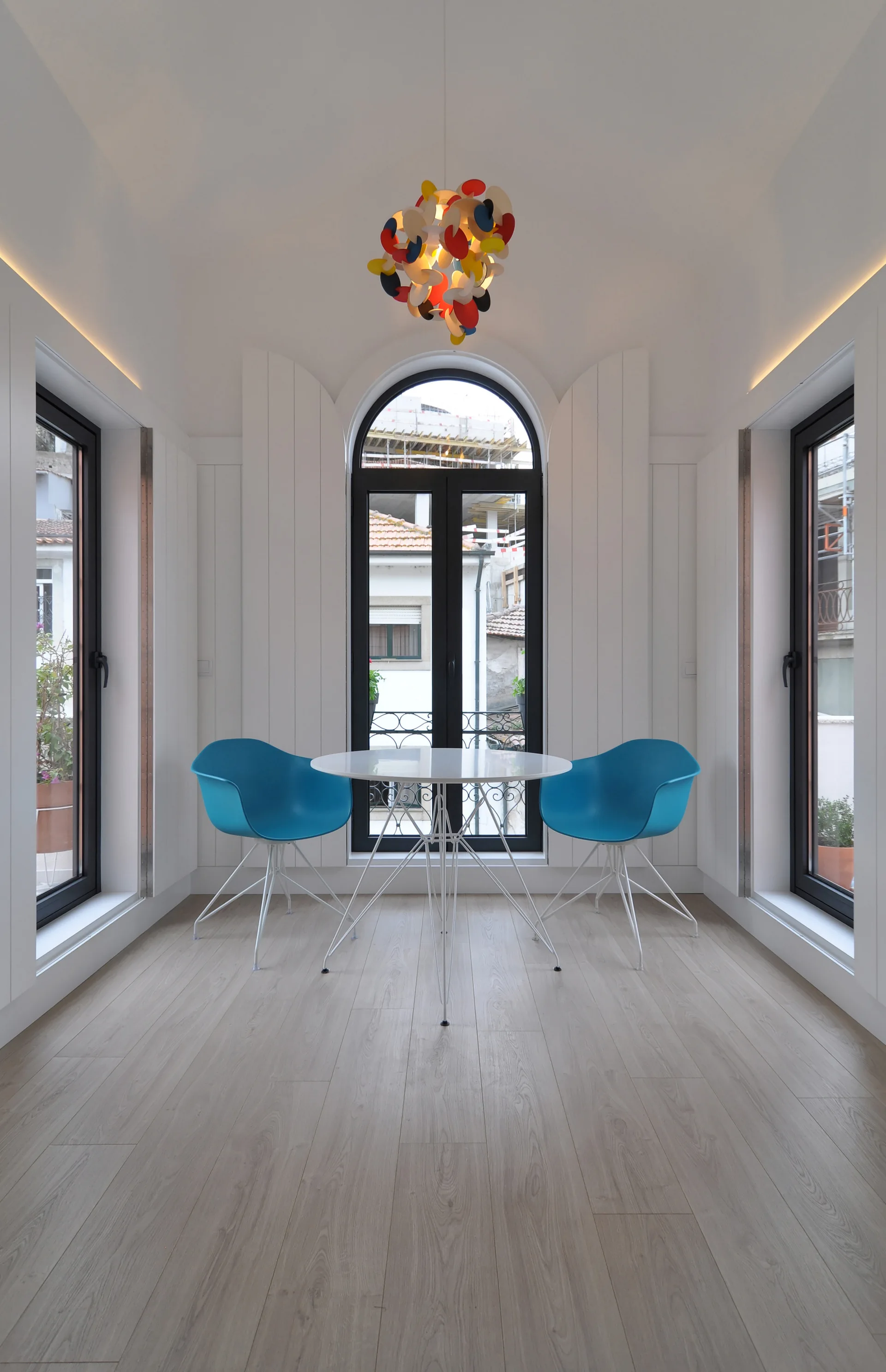 A bright dining area featuring a round white table, two blue plastic armchairs, and three black-framed windows under a multi-colored pendant light.