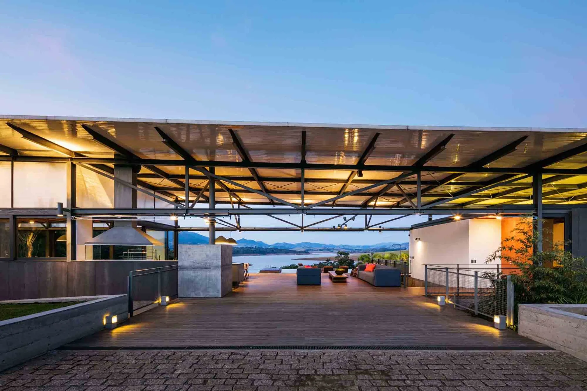 A covered wooden deck features a spacious seating area with grey sofas, a large steel roof, and a scenic lake view at dusk.
