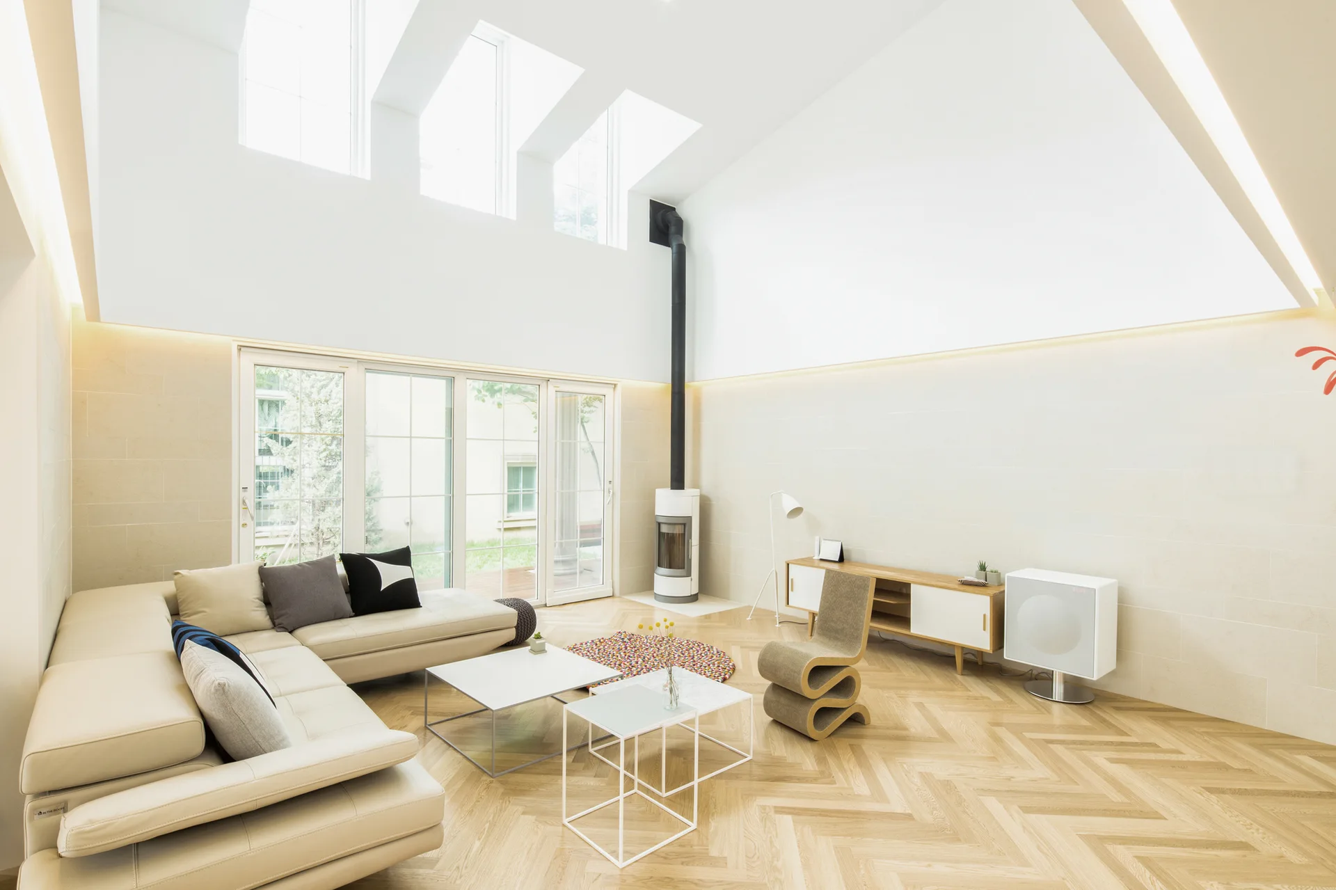 A bright living room features a cream sectional sofa, light wood herringbone flooring, a cylindrical white fireplace, and skylights in a high vaulted ceiling.