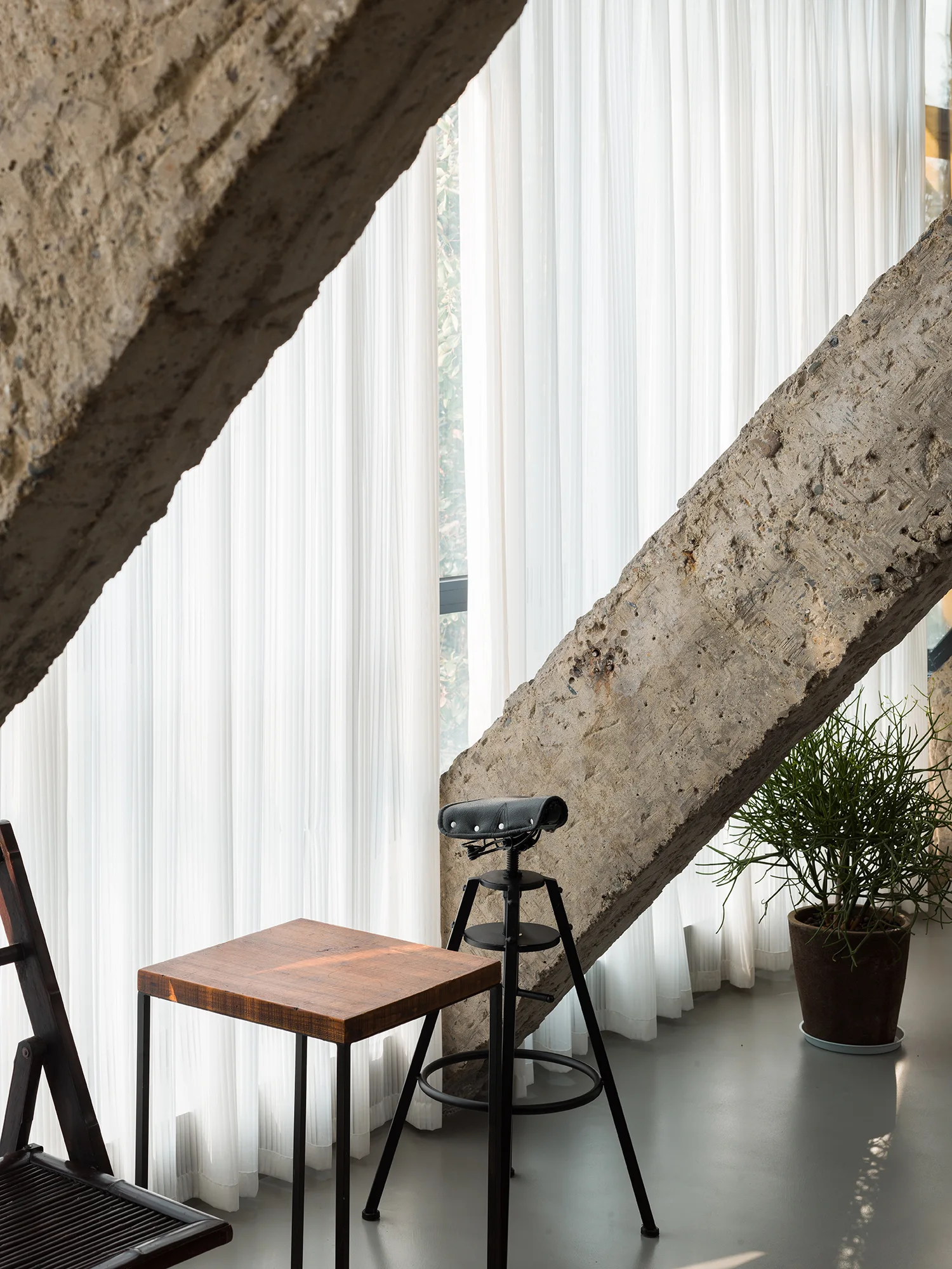 A minimalist room features a bicycle-seat stool and a wooden side table set against sheer white curtains and large diagonal concrete beams.
