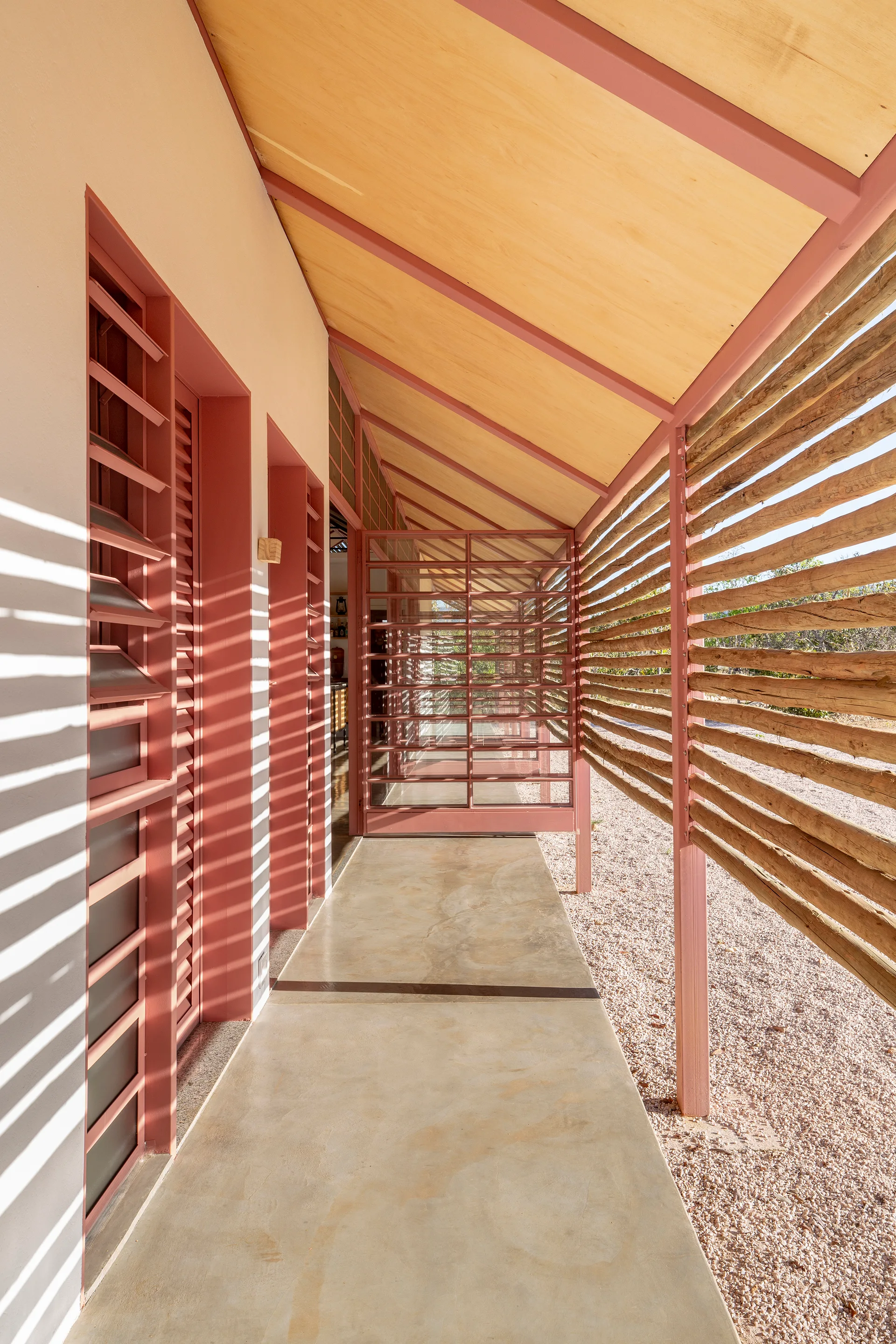 A concrete outdoor walkway features a dusty pink painted metal frame, a horizontal timber log screen, and louvered shutters casting direct shadows.