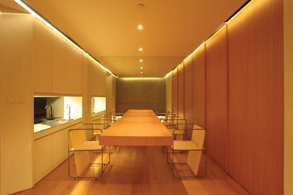 A long wood dining table sits centered in a narrow room with warm wood-paneled walls, built-in cabinetry, and glowing linear ceiling lights.