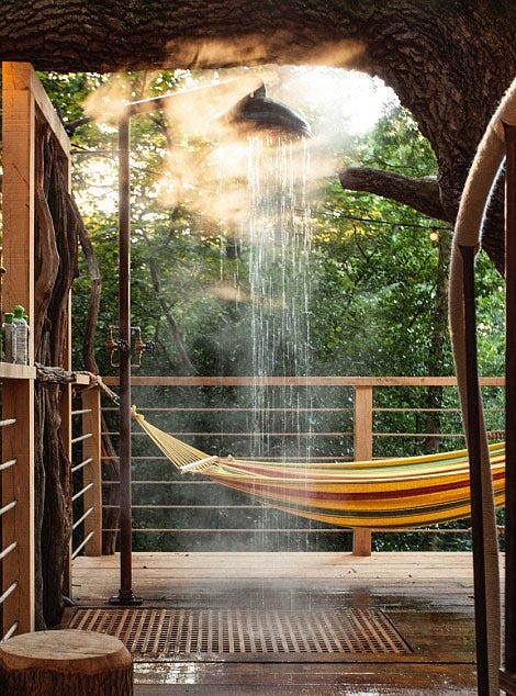 Water sprays from an outdoor shower fixture on a rustic wooden deck beneath a large tree canopy, with a colorful striped hammock hanging in the background.