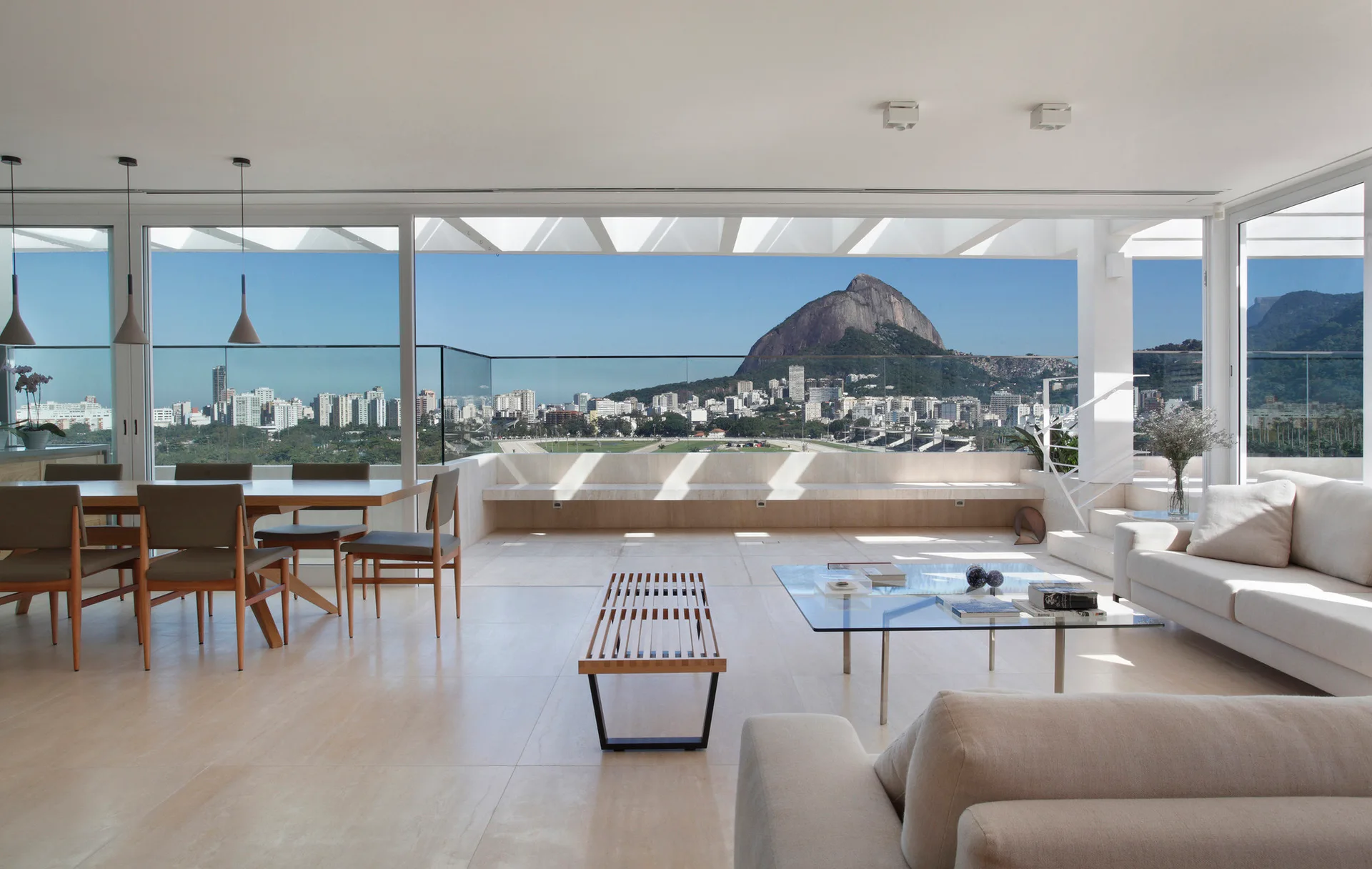 A bright open-plan living and dining space features white sofas, a wooden table, and expansive glass doors framing a city and mountain backdrop.