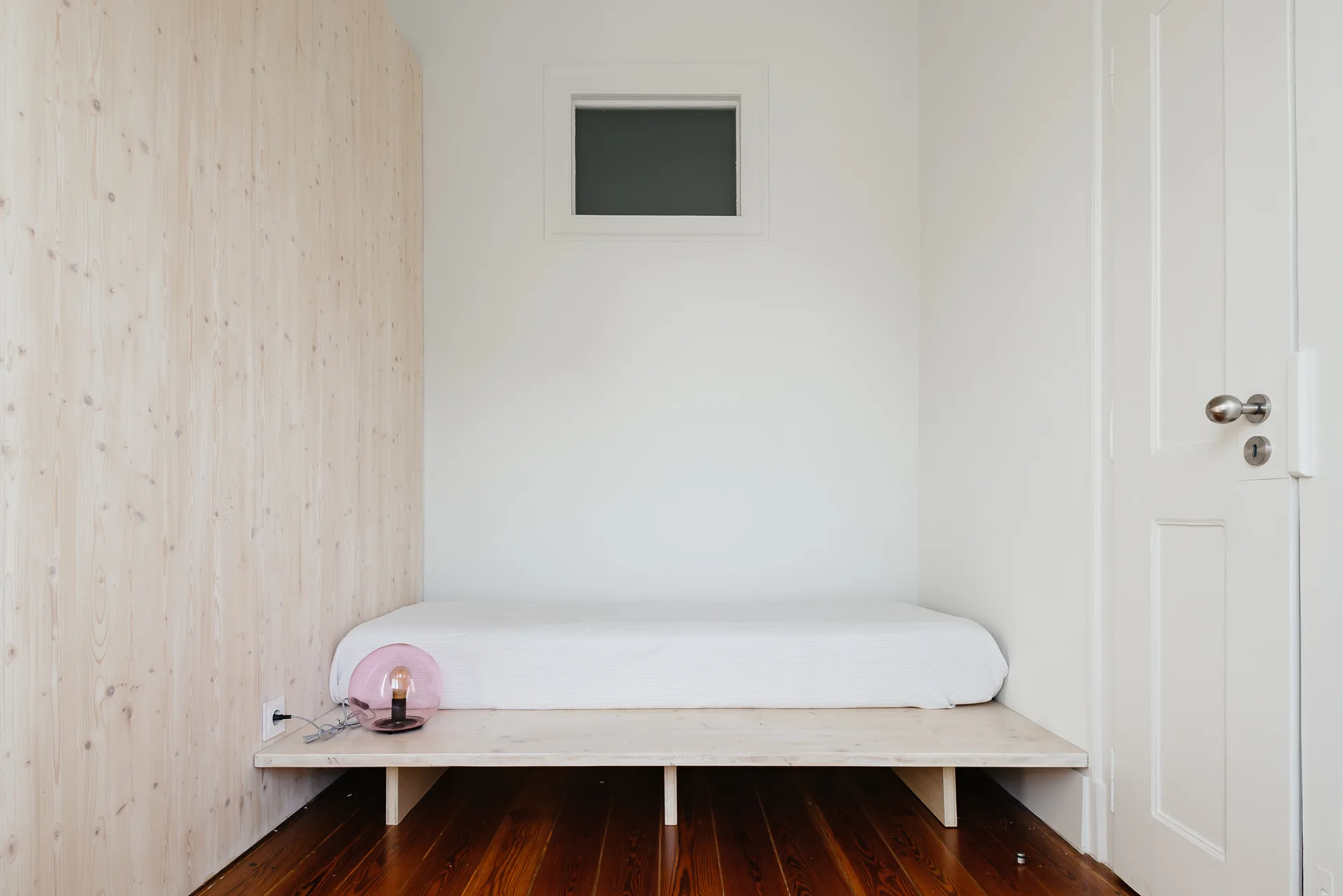 A minimalist sleeping nook featuring a white mattress on a low wooden platform bed, light wood wall paneling, and a small square window.