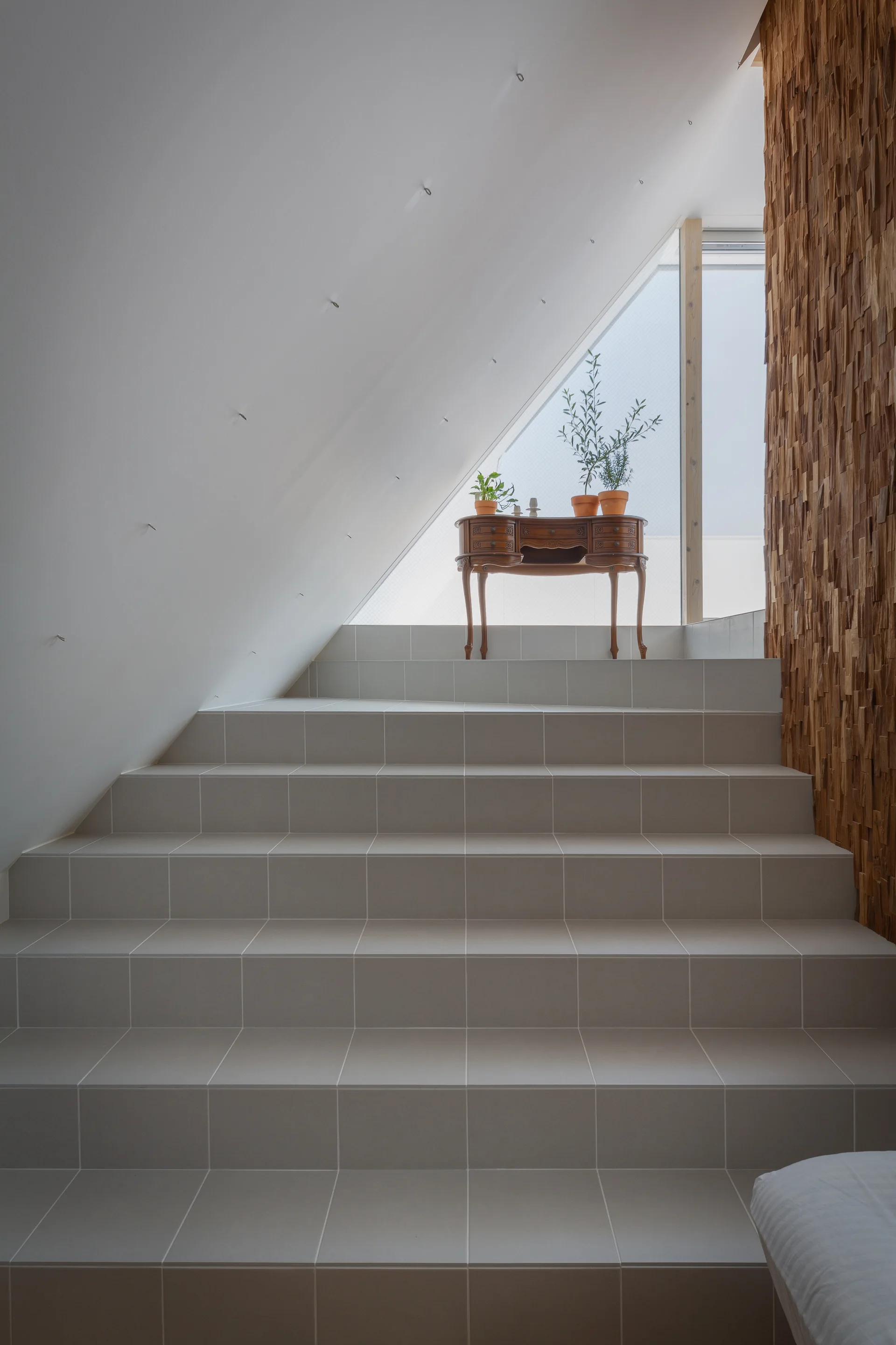 Wide tiled staircase leading to a bright landing with a vintage wooden console table, a sloped white ceiling, and a textured wood wall.