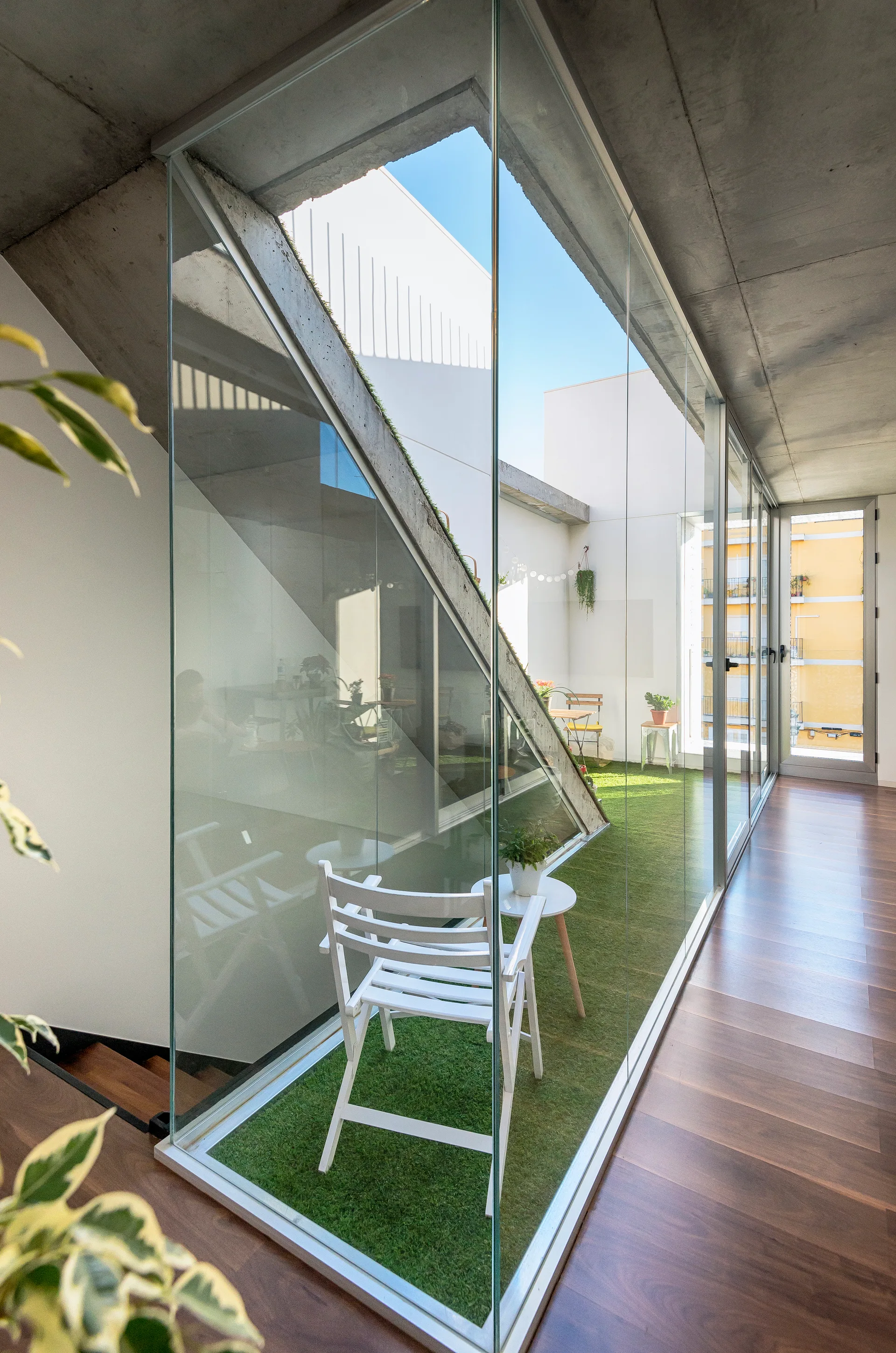 A floor-to-ceiling glass wall separates a wood-floored corridor from an enclosed courtyard featuring green turf, a white folding chair, and a concrete ceiling.