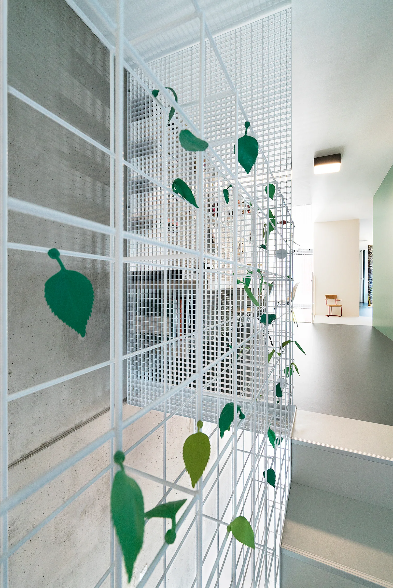 A minimalist hallway features a white metal mesh partition screen decorated with green leaves next to an exposed concrete wall and stairs.