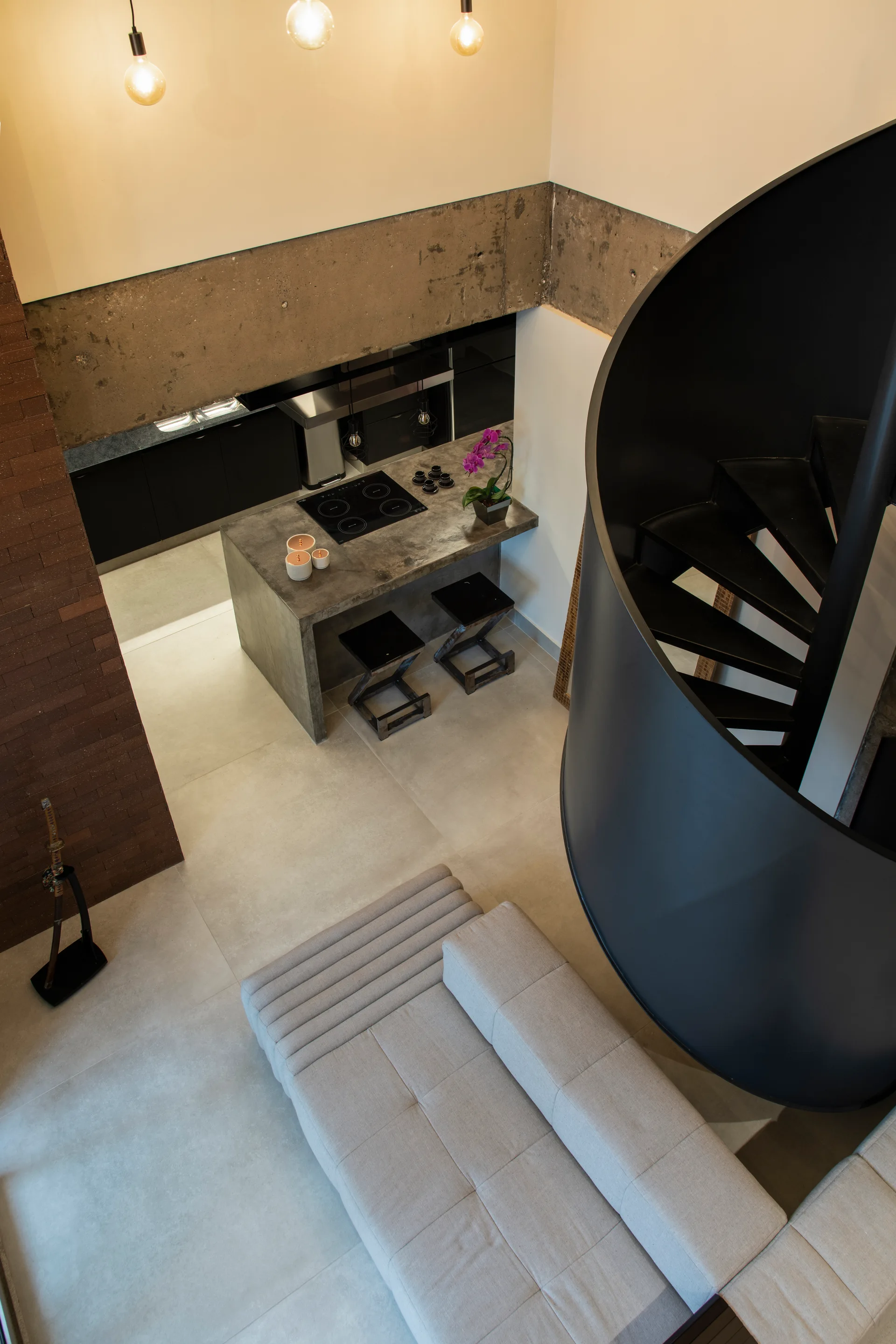 High-angle view of an industrial loft featuring a black spiral staircase, a grey modular sofa, and a concrete kitchen island.