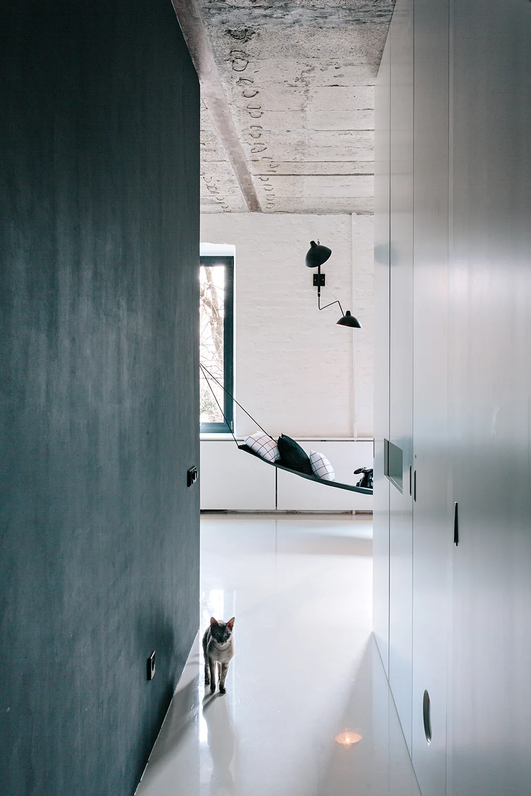 A minimalist hallway features seamless white cabinetry, an exposed concrete ceiling, and a highly reflective floor leading to a room with a black hammock.