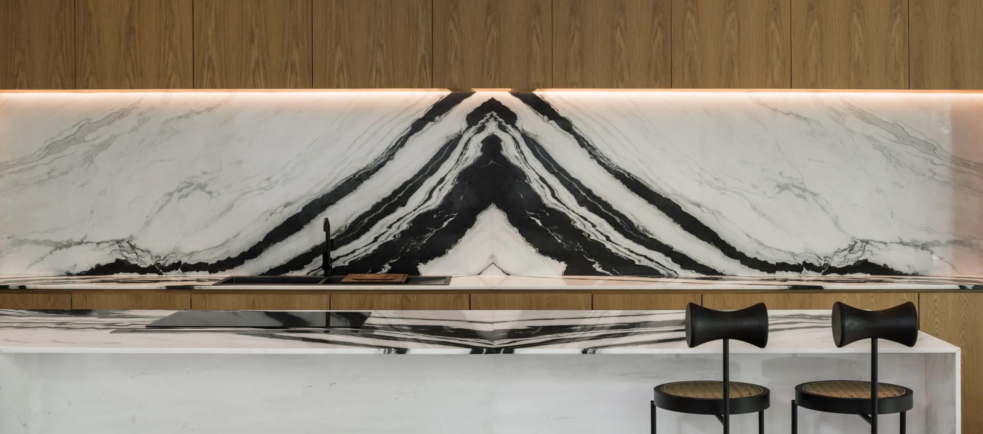 A modern kitchen featuring a bookmatched black and white marble backsplash, flat-front wood cabinets with under-cabinet lighting, and a matching island.