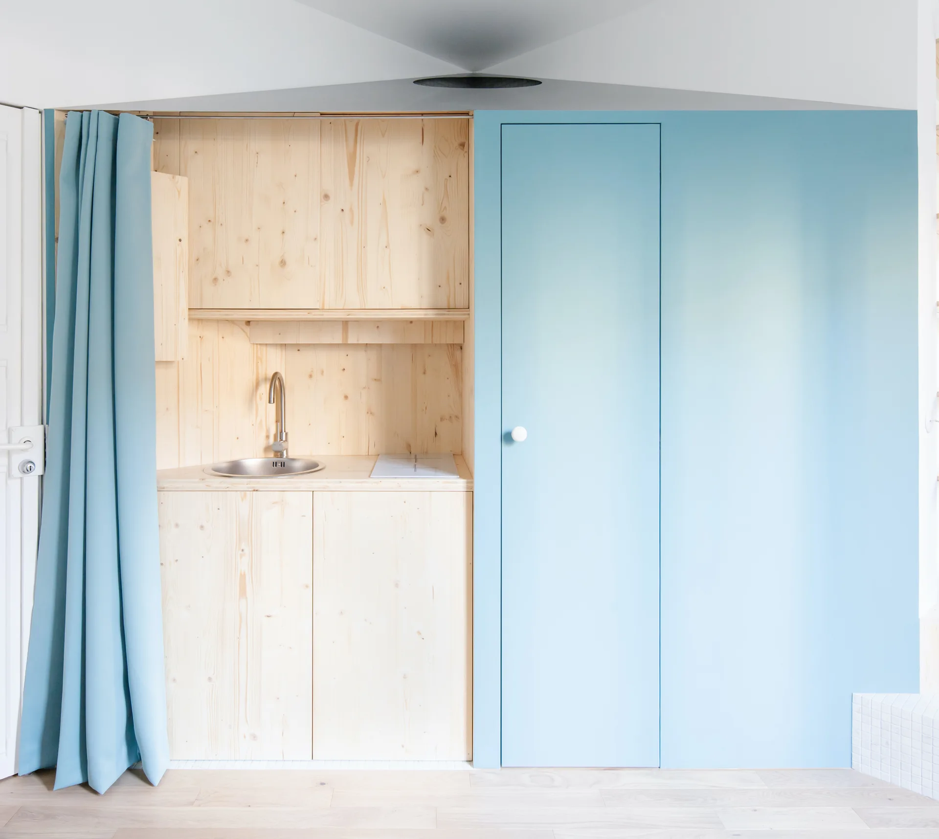 A compact light wood kitchenette with a small steel sink is set next to a solid light blue wall panel and a matching blue fabric curtain.