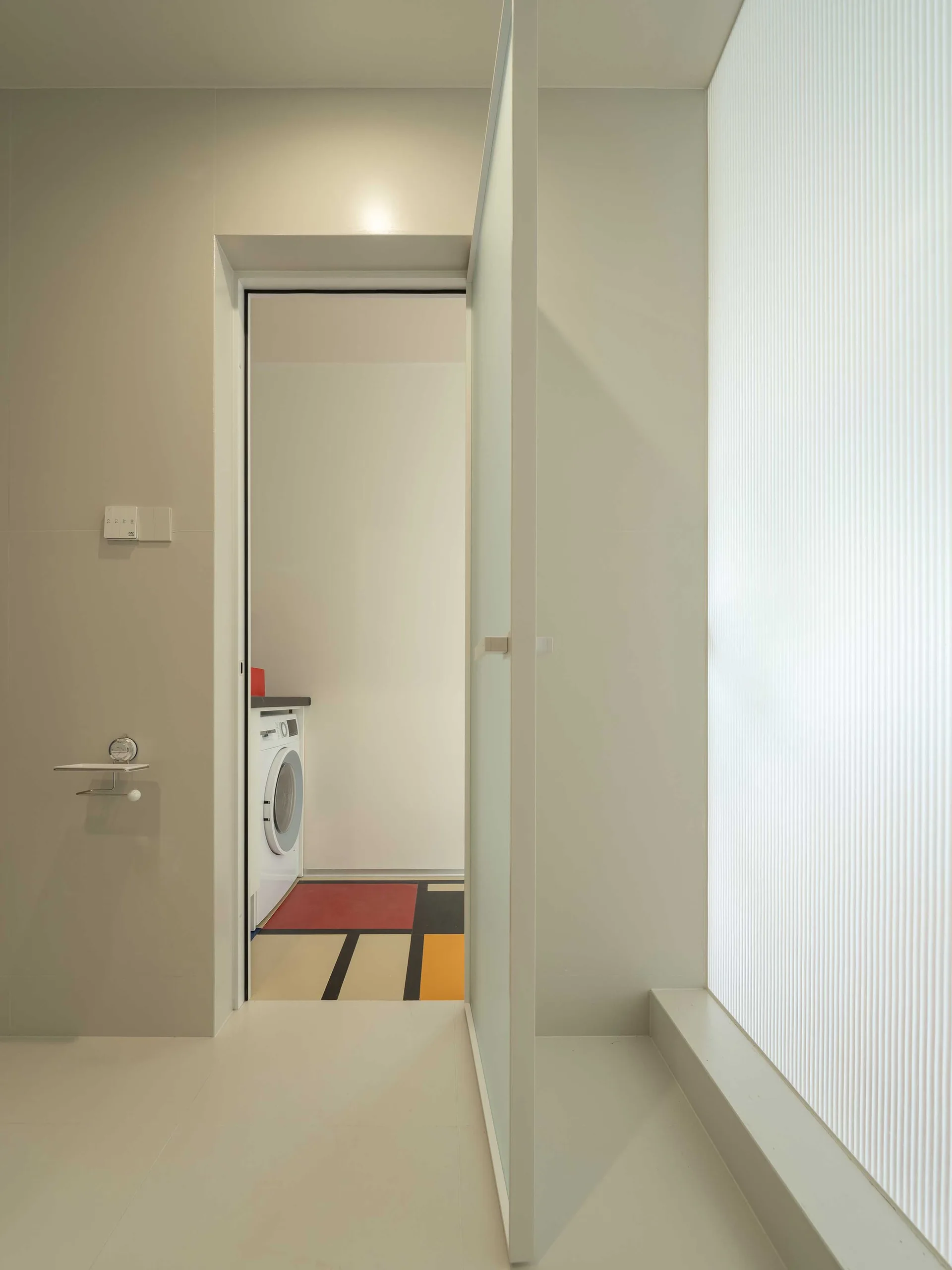 A frosted glass door opens to a minimalist laundry room featuring a geometric, color-blocked tile floor and a white washing machine.