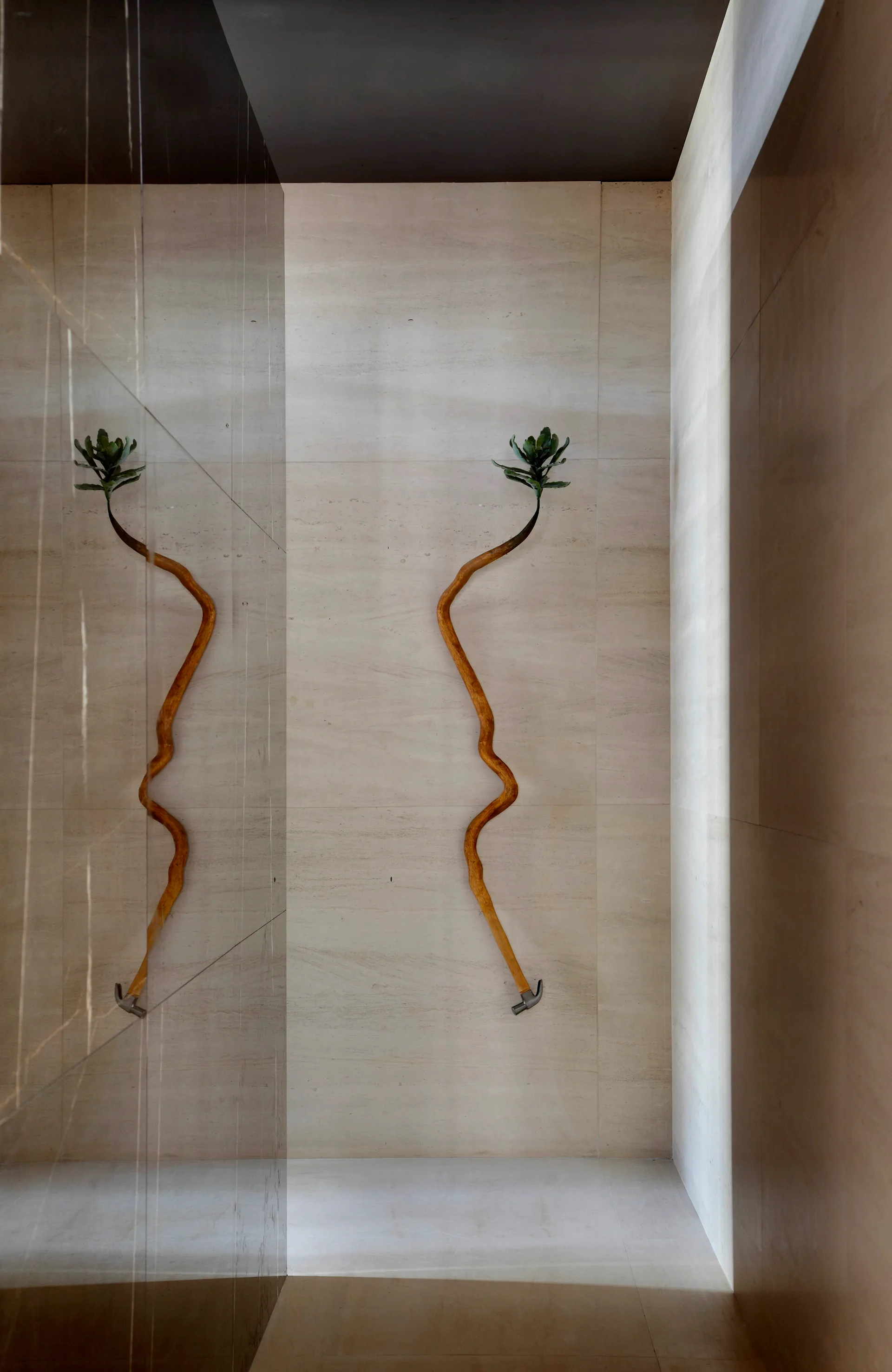A curved wooden branch sculpture topped with a small succulent hangs on a beige stone tile wall next to a large mirror panel.