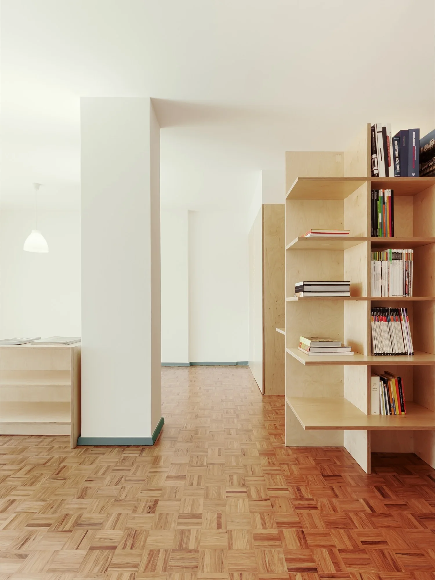 A minimalist interior room featuring a light plywood bookshelf, parquet wood flooring, and a large white structural column with blue baseboard trim.