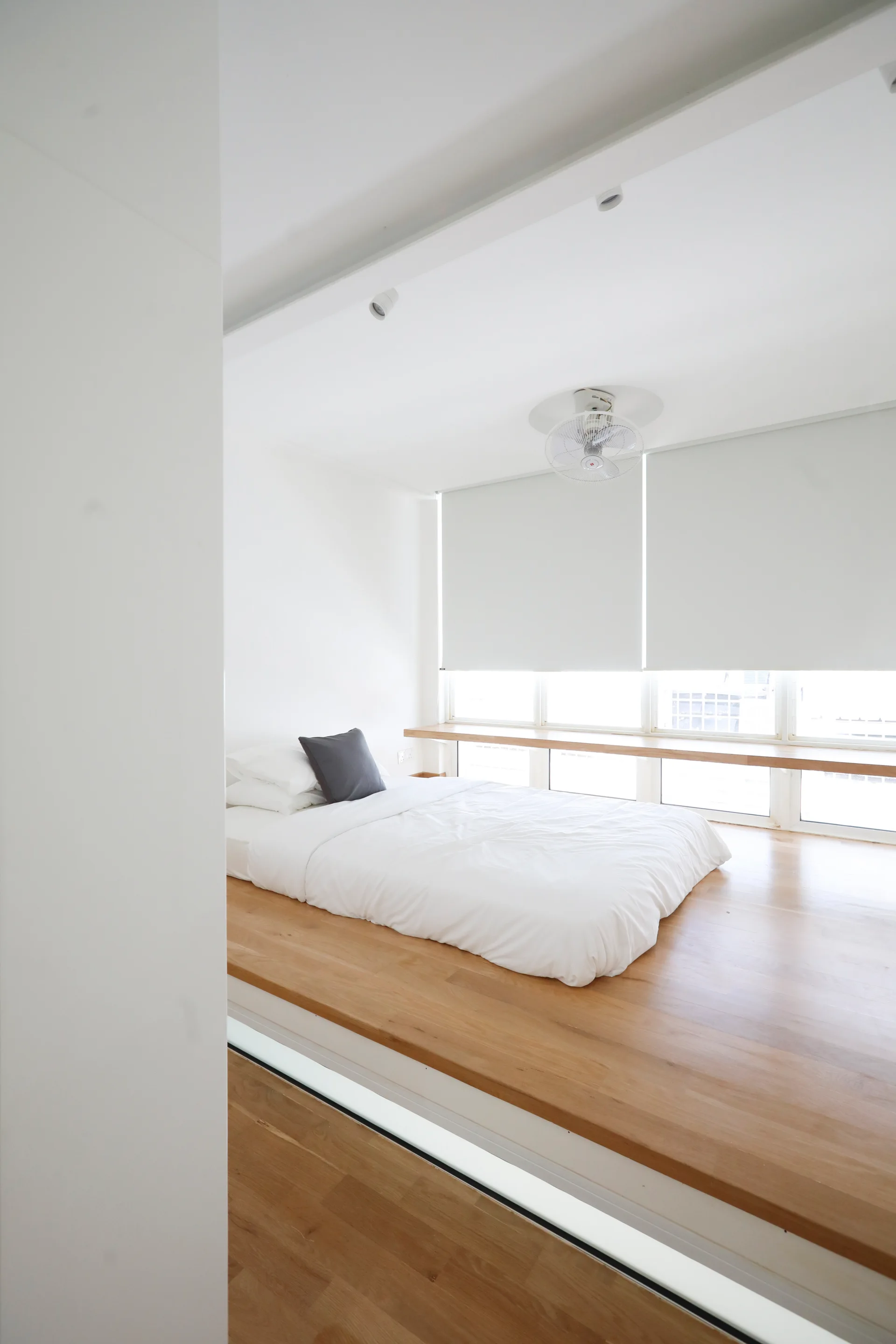 A bed with white linens sits on an elevated wood platform in a minimalist bedroom featuring a long window desk ledge.