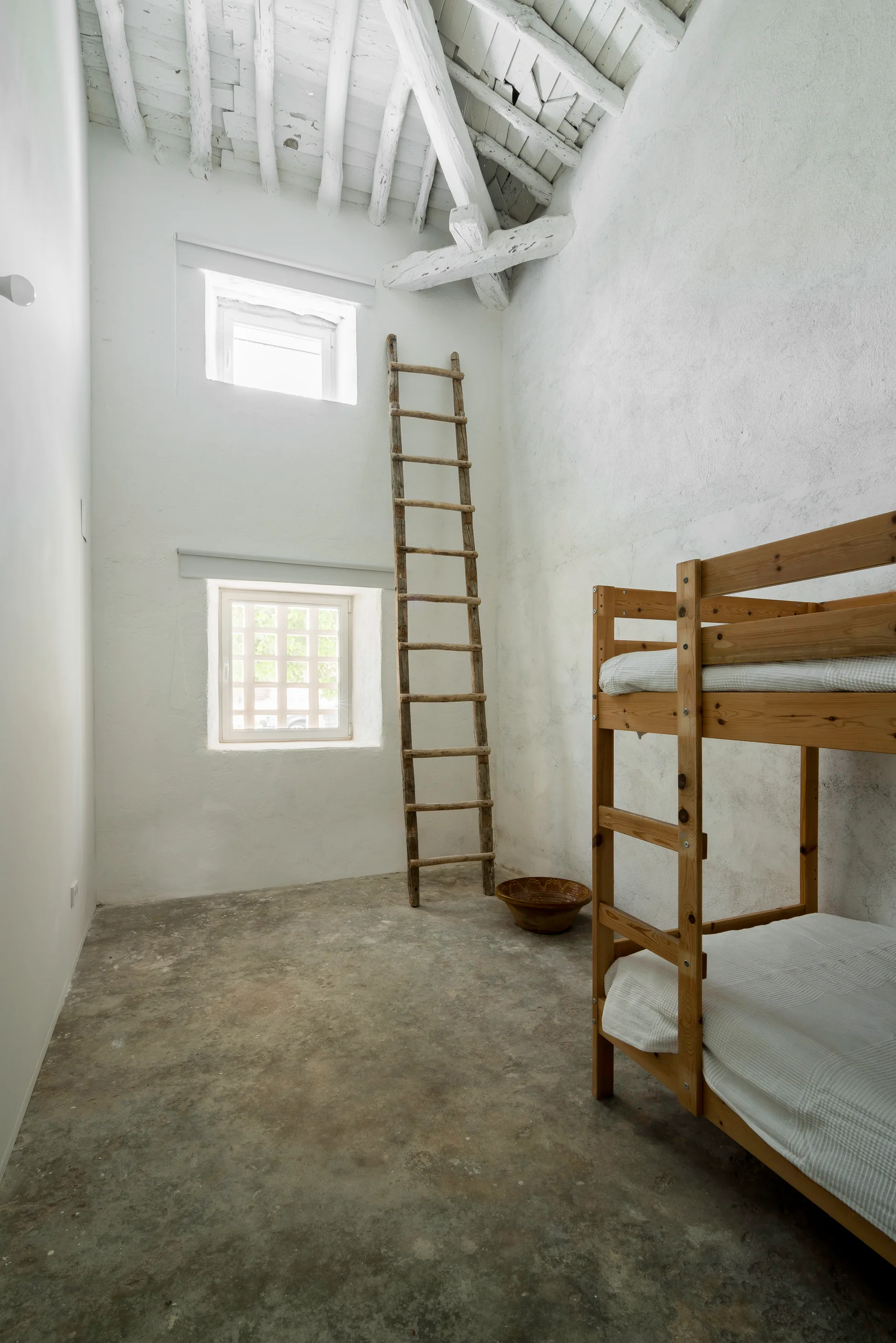 A rustic bedroom with white stucco walls, a grey concrete floor, a wooden bunk bed, and a tall wooden ladder leaning near stacked square windows.