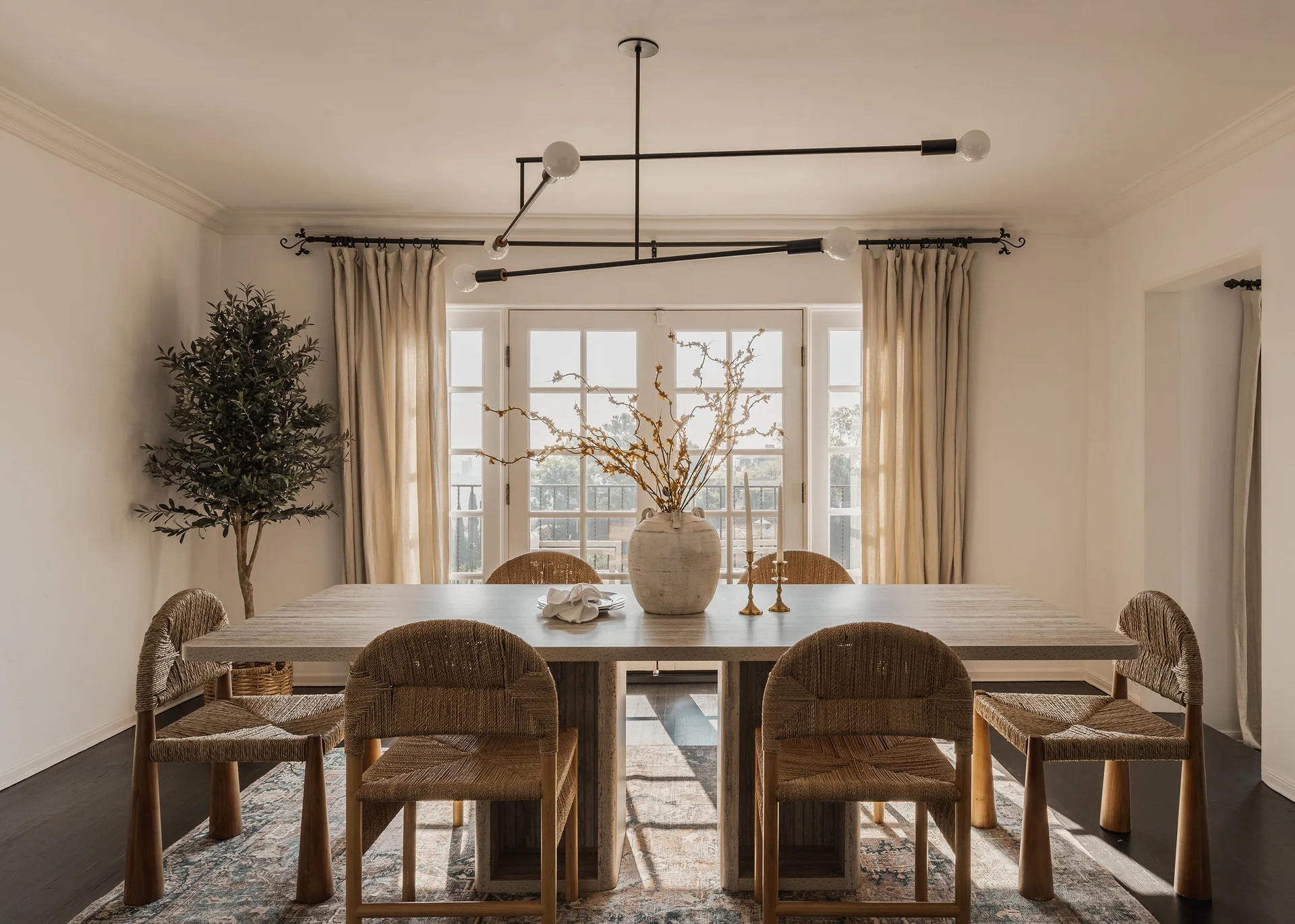 A dining room features a stone-topped table surrounded by woven chairs on a patterned rug under a modern black linear chandelier.