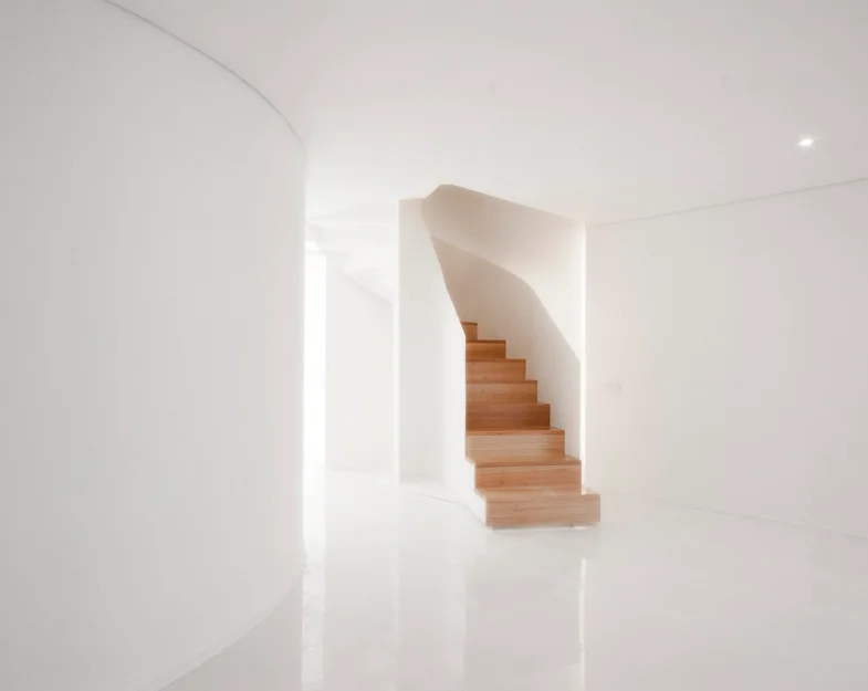 A minimalist white entryway featuring a curved wall, a highly reflective glossy floor, and a central staircase with warm wooden treads.