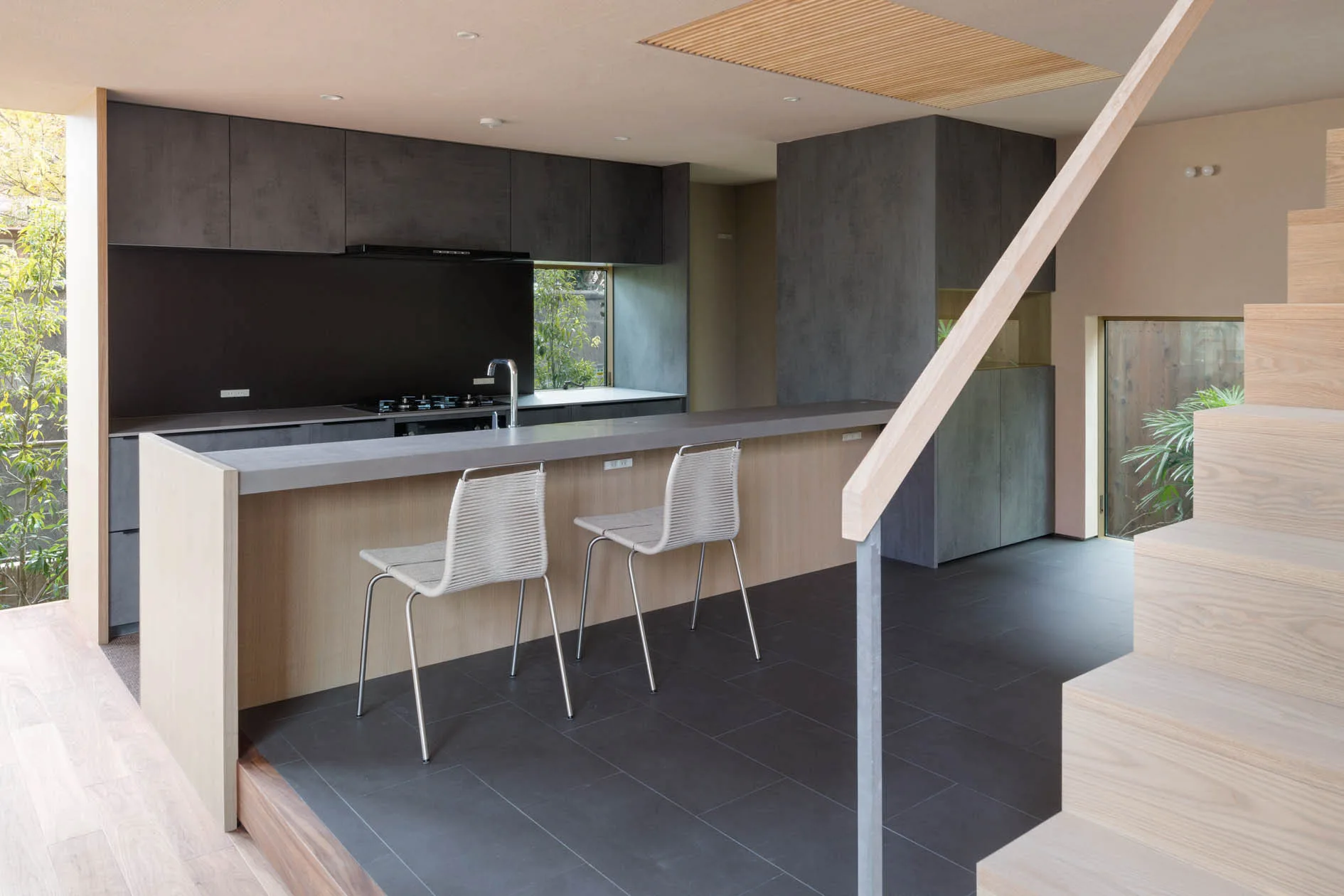 A minimalist kitchen features charcoal grey cabinetry, a light wood island with two woven stools, and a wooden staircase in the foreground.