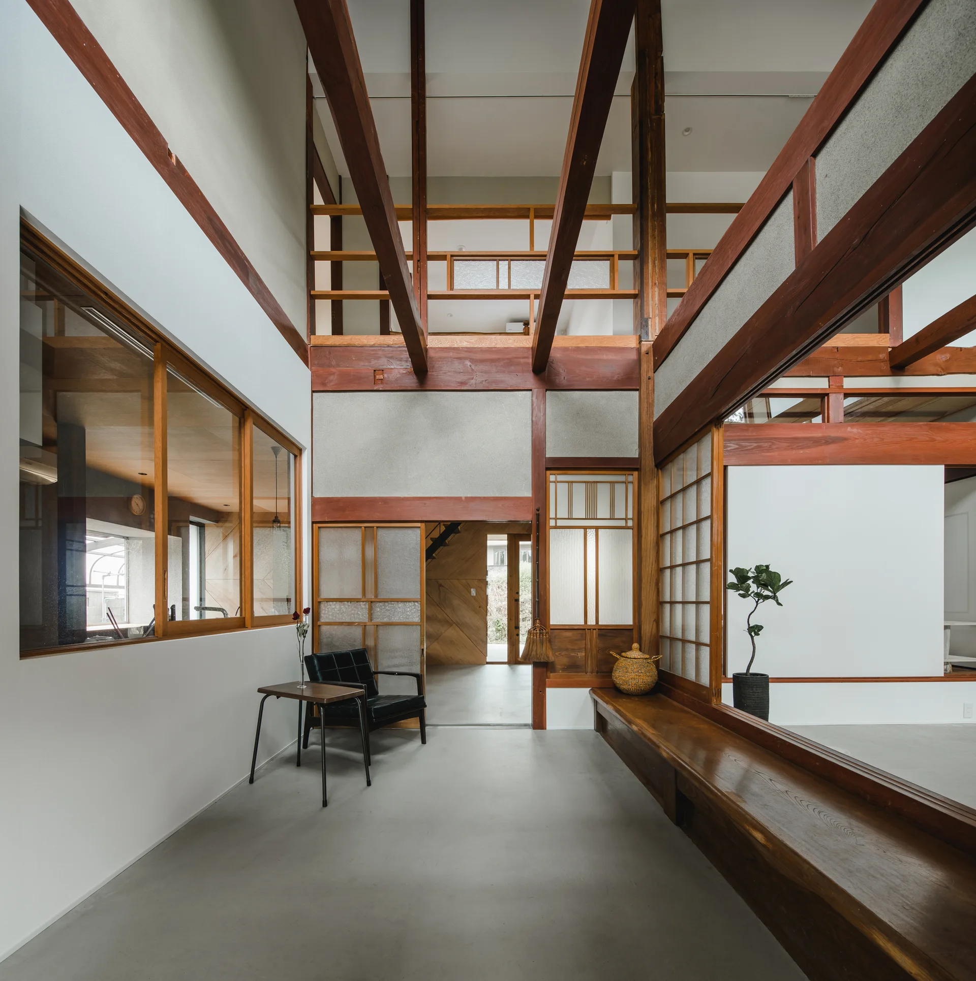 A spacious double-height entryway featuring exposed dark wood beams, a concrete-like floor, a long built-in bench, and traditional Japanese shoji screens.