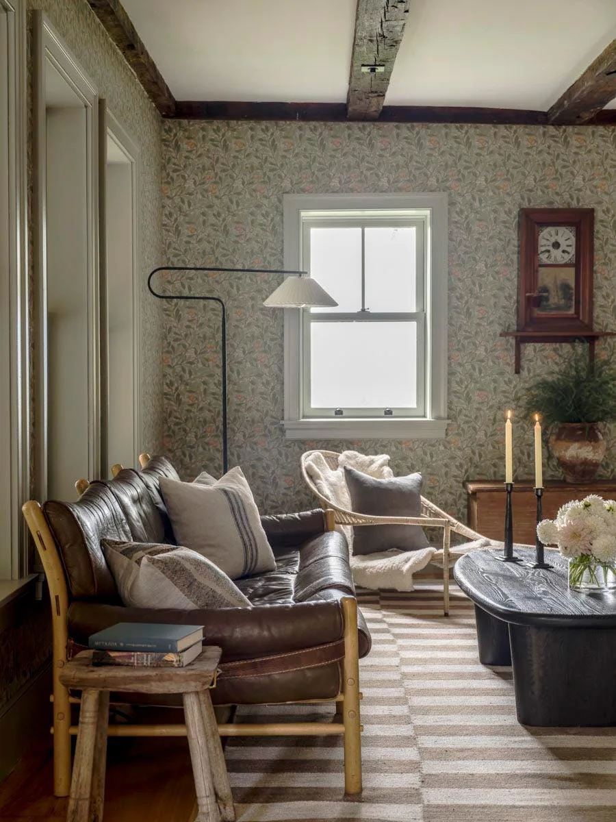 A rustic living room with a brown leather sofa, a wicker armchair, green floral wallpaper, and dark exposed ceiling beams.