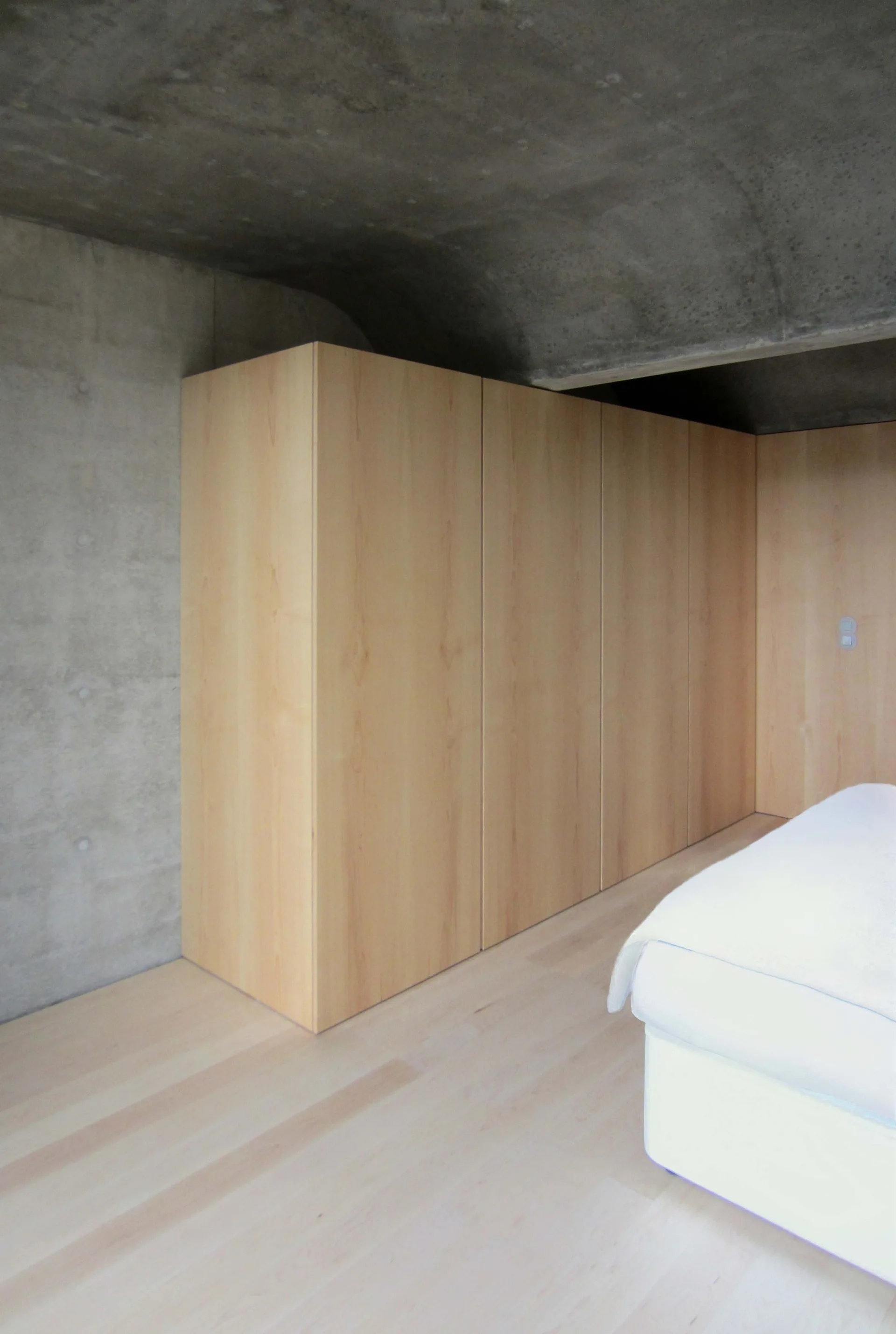 A minimalist bedroom features a built-in light wood wardrobe matching the wood floor beneath a curved, exposed concrete ceiling.
