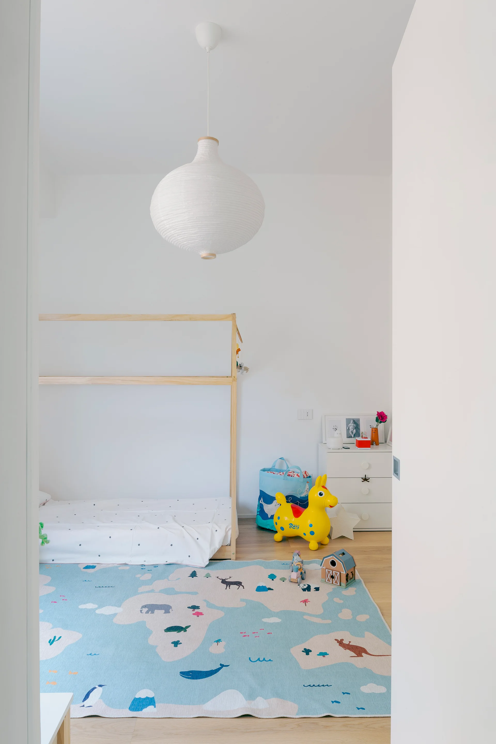 A bright minimalist kids bedroom features a wooden house bed frame, a light blue world map rug, white walls, and a paper pendant light.