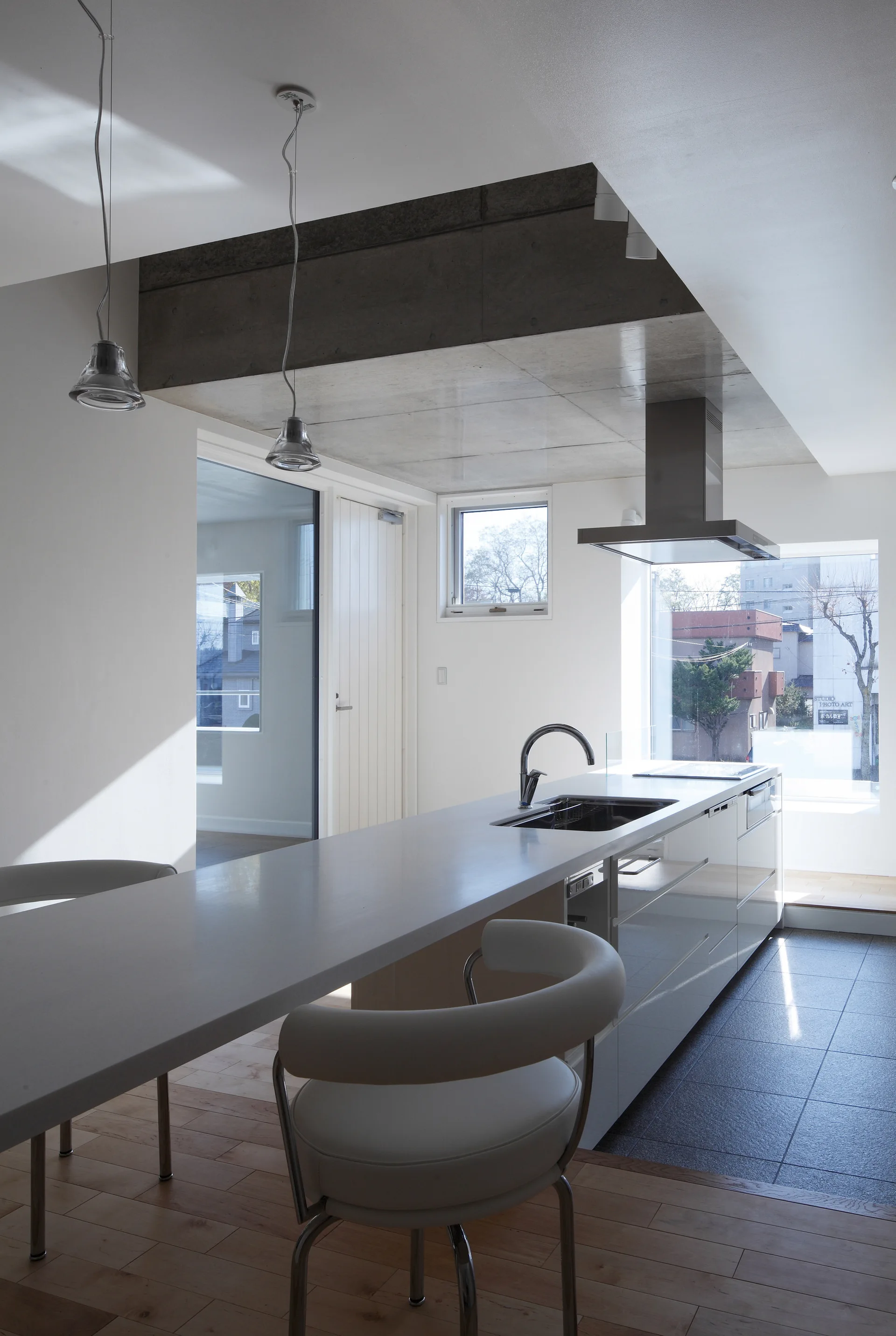 A modern minimalist kitchen featuring a long white island with a sink, white bar stools, and a dropped concrete ceiling section.