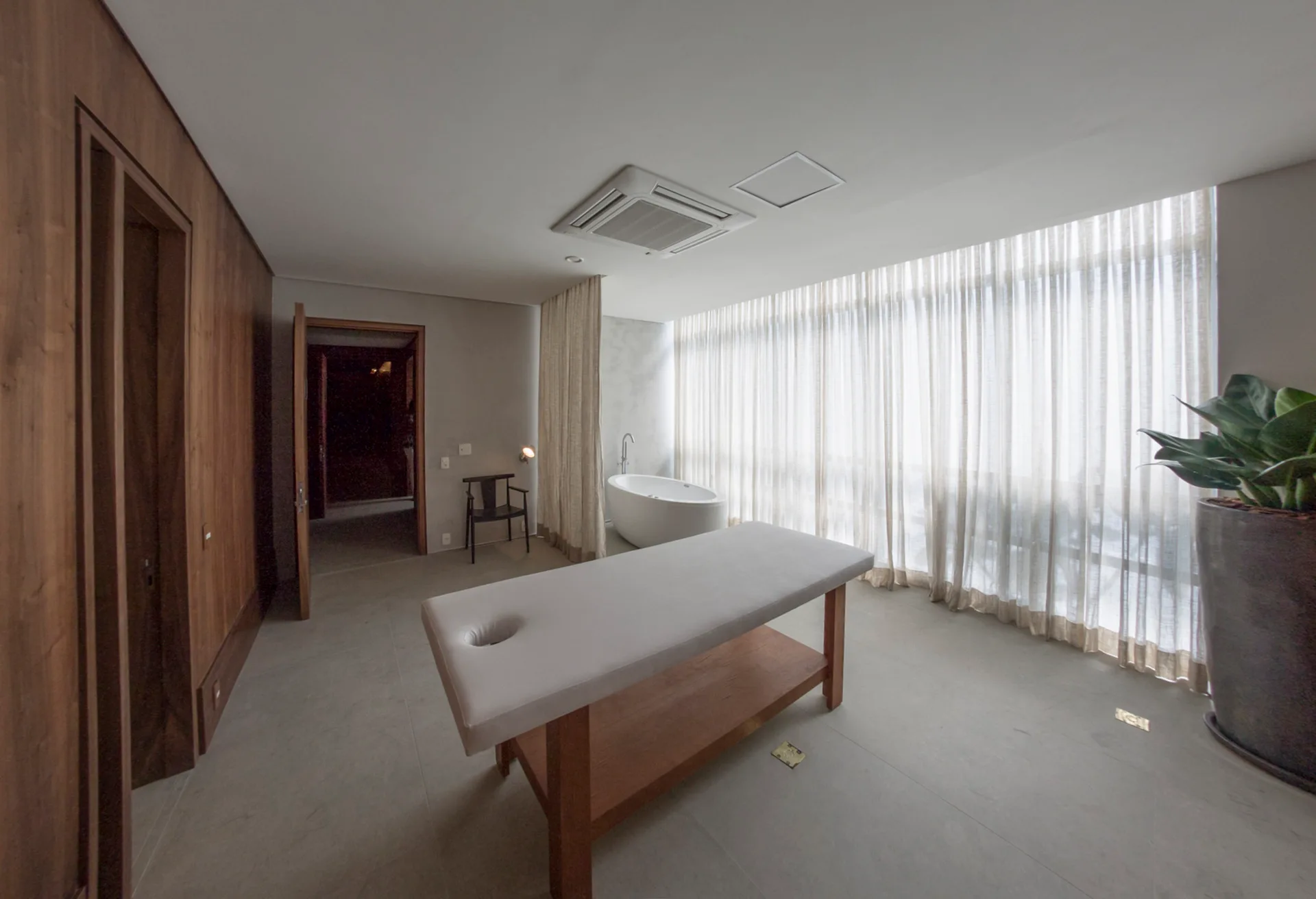 A minimalist wellness room featuring a wooden massage table, a white freestanding bathtub by sheer curtains, and warm wood wall paneling.