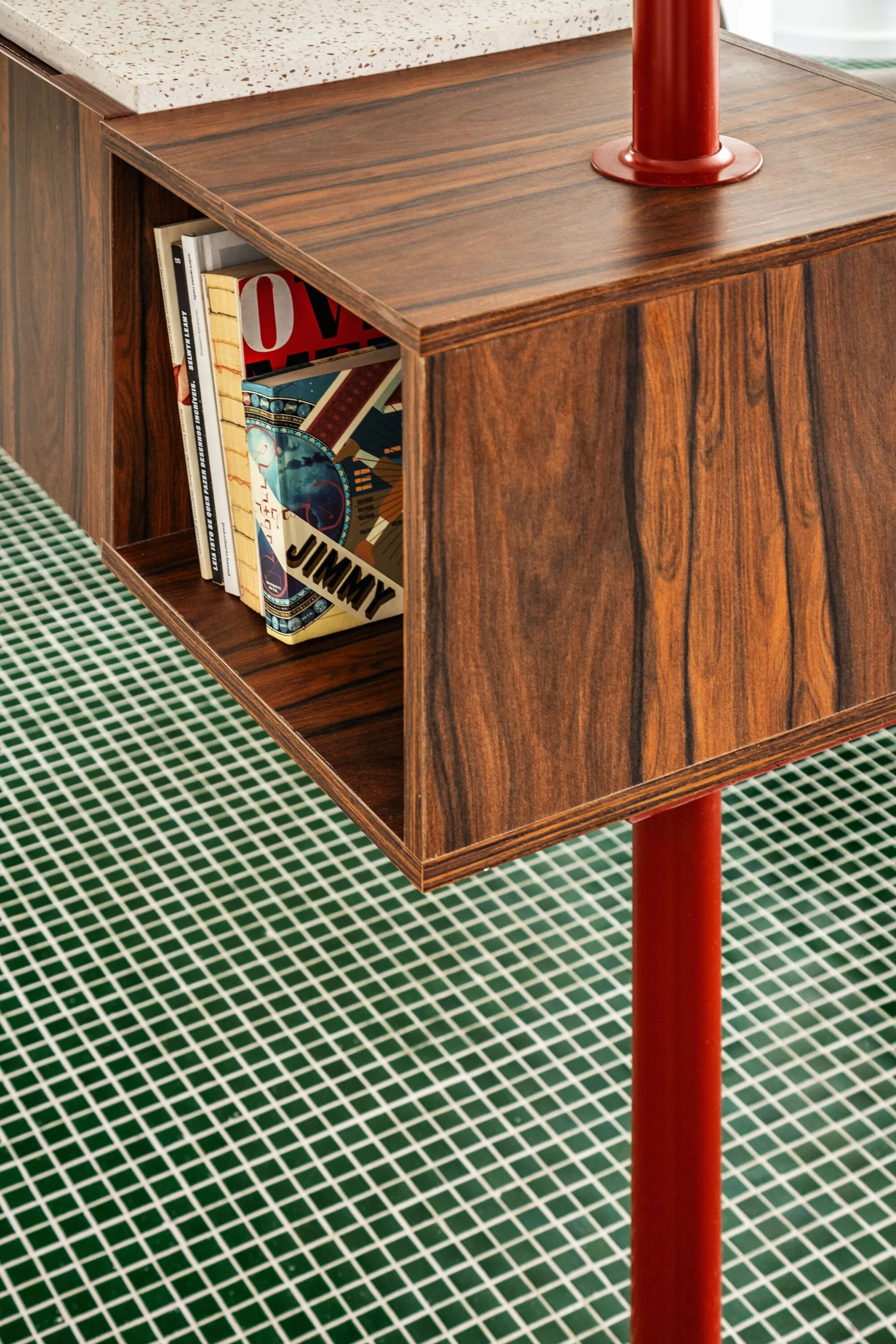 A wood-grained floating cabinet with a terrazzo top is supported by a red metal post above a green tiled floor.