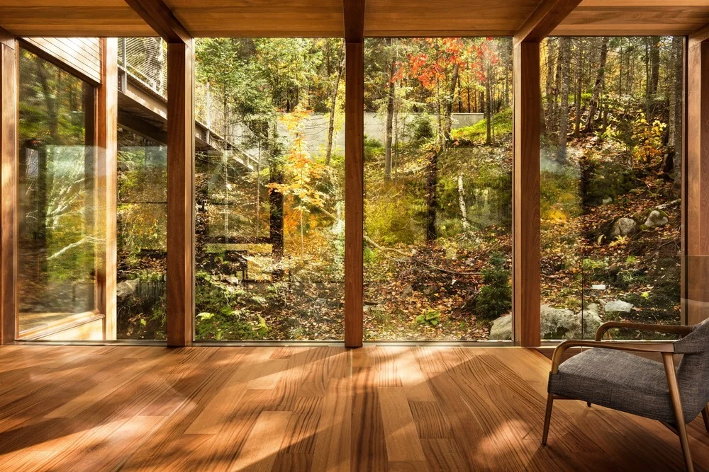 A minimalist room with polished wood flooring and wood-framed floor-to-ceiling windows looking out onto a dense autumn forest.