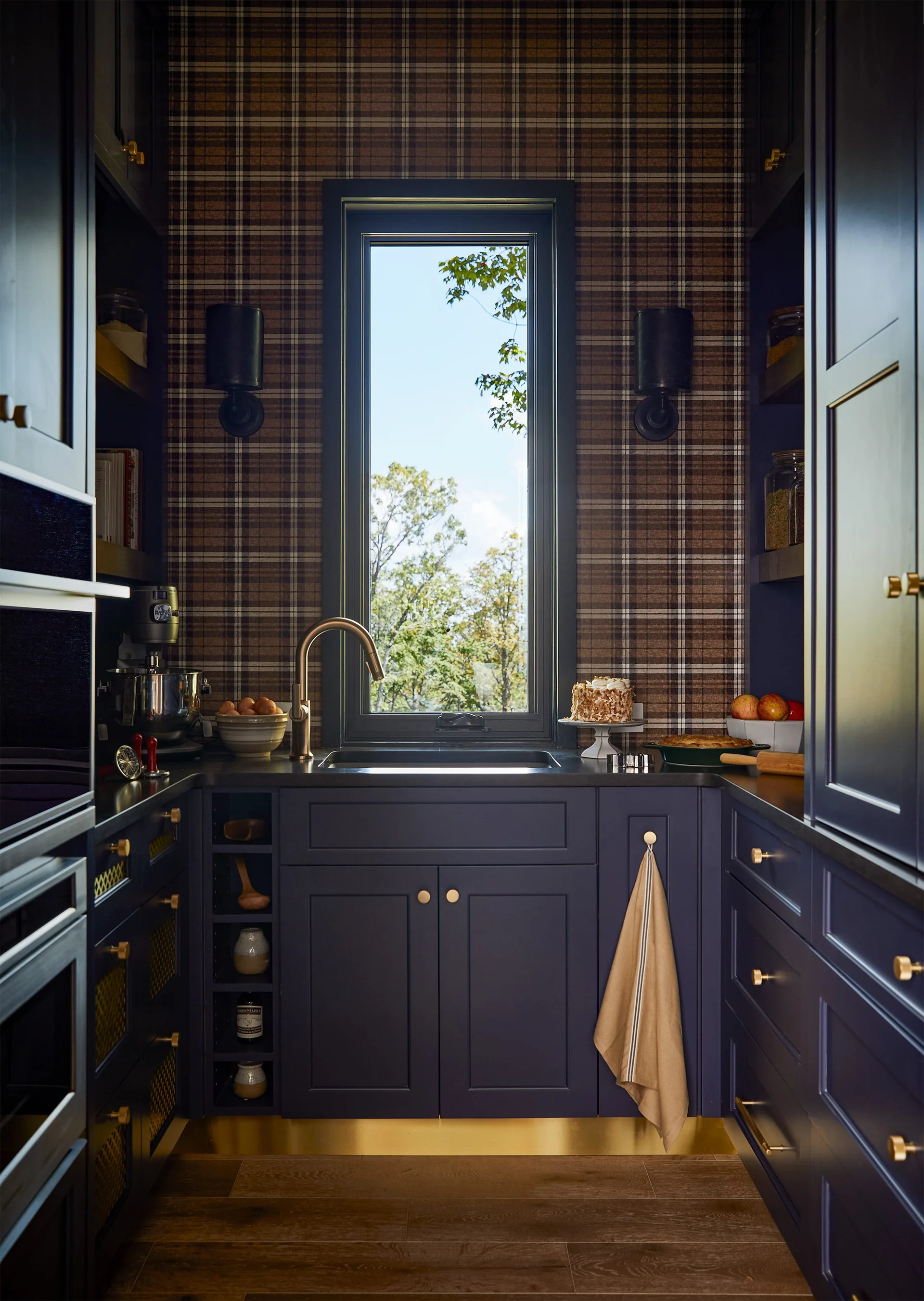 A dark navy kitchen nook with shaker cabinets, a central vertical window, brass hardware, and brown plaid wallpaper.